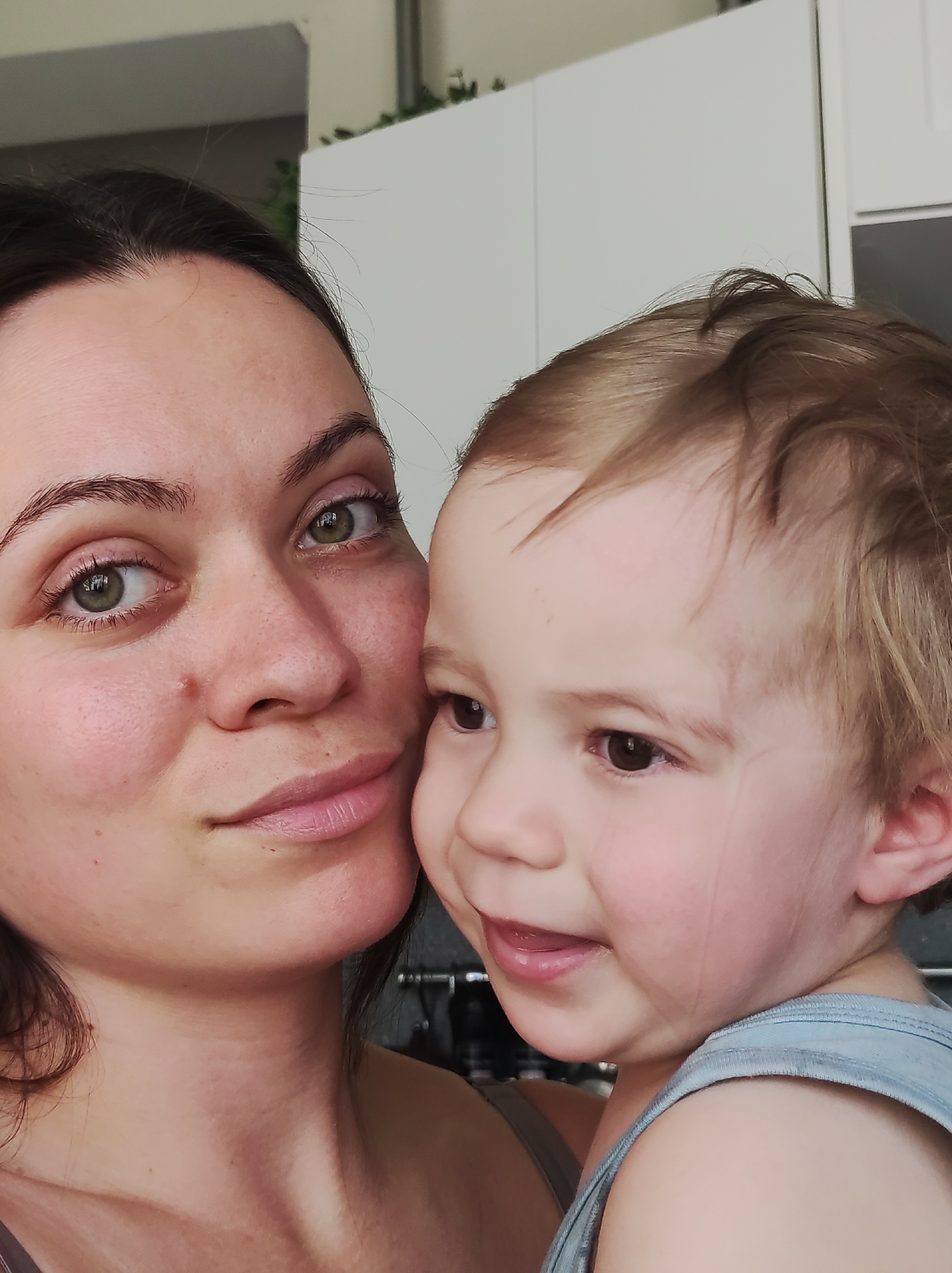 A fair-skinned woman with dark hair and green eyes is holding a young child with fair skin and light brown hair. The woman is looking at the camera with a slight smile, while the child is looking to the side with a smile and their mouth slightly open. Both are wearing light-colored tops.
The background includes a white kitchen cabinet and a stainless steel bar. The image appears to be taken indoors in a residential setting. There is no information provided in the image to connect it specifically to the city of Chaikovsky, Russia.