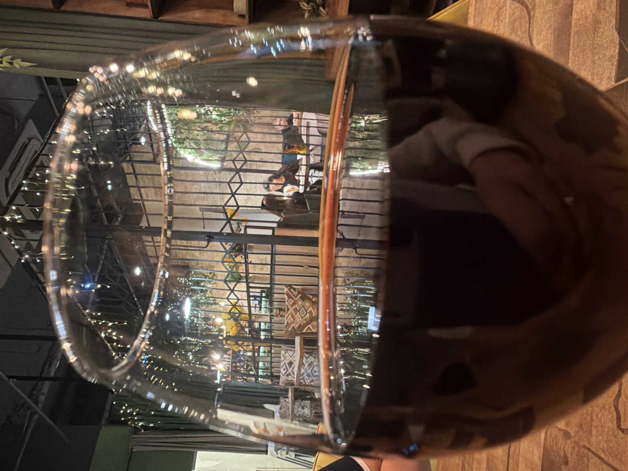 The image depicts a close-up of a wine glass filled with a dark liquid, likely red wine, with a distorted view of the surroundings visible through the glass. The backdrop appears to be a restaurant or outdoor seating area, possibly in the evening, with string lights and decorative elements. Tables, chairs, and people are discernible through the glass's refraction. The location is speculated to be Izhevsk, Russia. The focus is primarily on the wine glass and the reflections it captures, blurring the environmental context. The perspective is somewhat unusual, suggesting the image was taken from a low angle.