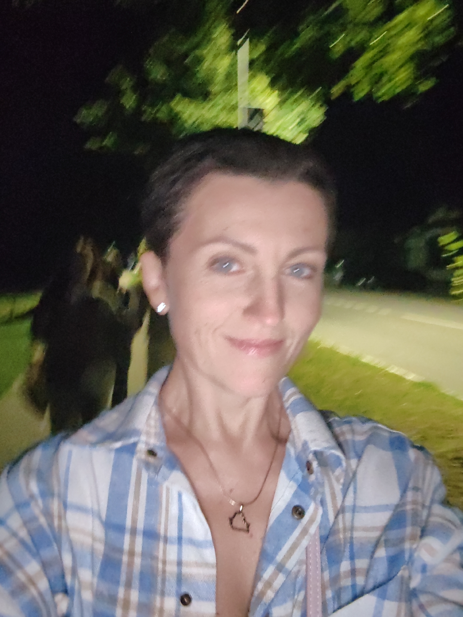 The image is a selfie of a woman with short brown hair and blue eyes, wearing a blue and white plaid shirt. She is wearing a necklace with a small triangular pendant. She is standing outdoors at night, with trees and a street visible in the background. The location is identified as the city of Myadzyel, Belarus. Several other people are visible in the background, blurred due to the low light conditions and movement. The lighting suggests that the photo was taken under artificial light sources, possibly streetlights or lights from nearby buildings.