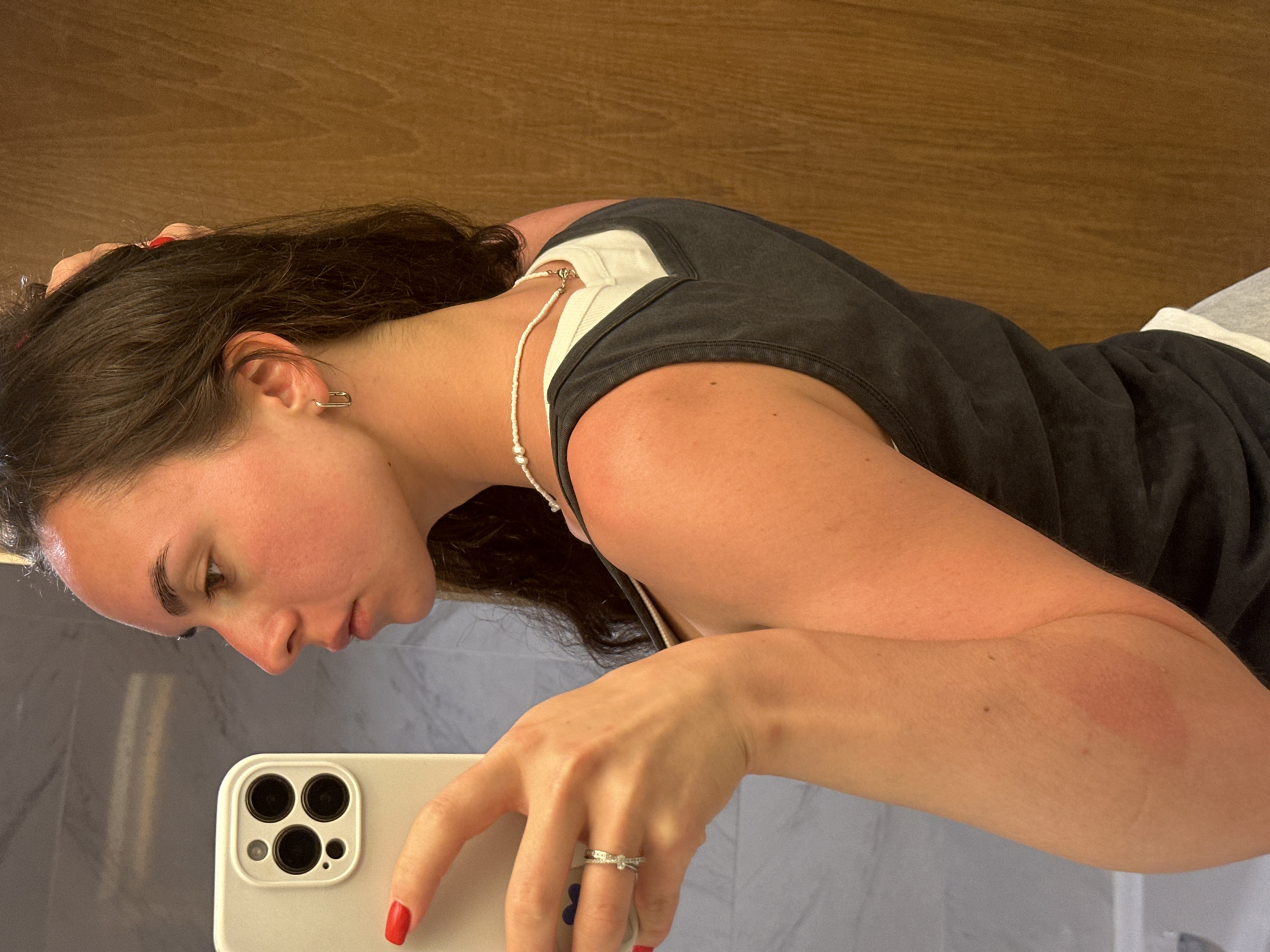 A woman with dark hair and fair skin is taking a selfie in a bathroom. She is wearing a dark grey tank top over a white shirt and a pearl necklace. She is holding a white smartphone with red nail polish on her fingers. A ring is visible on her left ring finger. The background includes a wooden wall and a light grey surface, possibly a countertop or sink. There is no indication of this being taken in Pattaya City, Thailand.