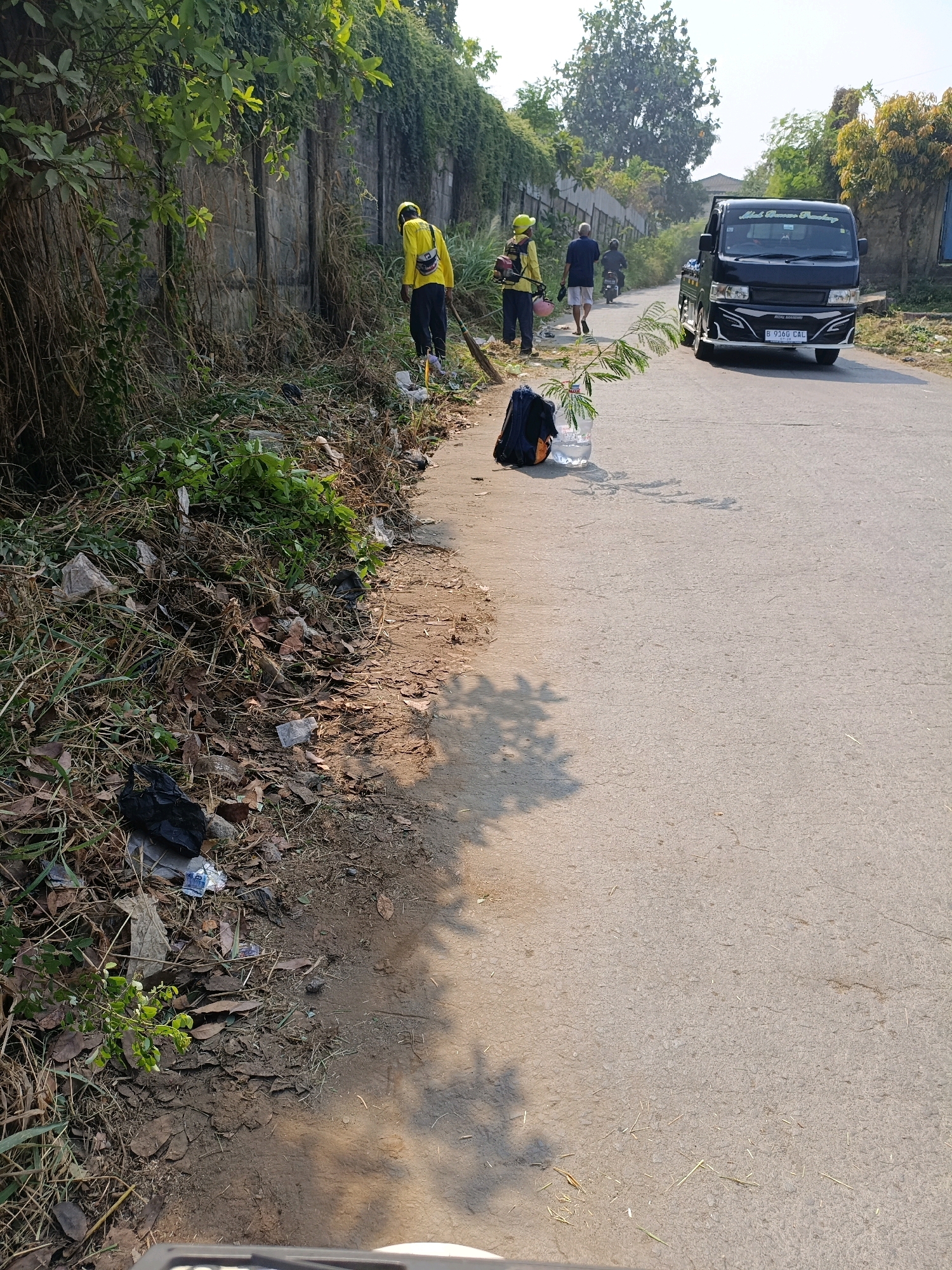 In Tangerang, Indonesia, a scene unfolds along a paved road with a concrete retaining wall on the left, overgrown with vegetation. The foreground is a verge of dry grass, fallen leaves, and scattered refuse including plastic bags and debris. Several individuals are engaged in clearing the roadside.
One person, wearing a yellow shirt, yellow hard hat, and dark pants, is sweeping the ground with a broom. Next to them, another individual in a yellow shirt and dark pants holds a pink bucket and a grass-trimming tool. Further down the road, two other individuals are walking, one wearing a dark blue shirt and shorts, the other on a motorcycle.
A black pick-up truck with a license plate reading "B 9160 CAL" is driving on the road, heading away from the viewer. A dark backpack is placed on the roadside next to a clear plastic bottle containing a small green plant. The overall environment suggests an outdoor, possibly semi-urban setting, with the activity focused on roadside maintenance or cleanup. The lighting indicates it is daytime, with sunlight casting shadows.
