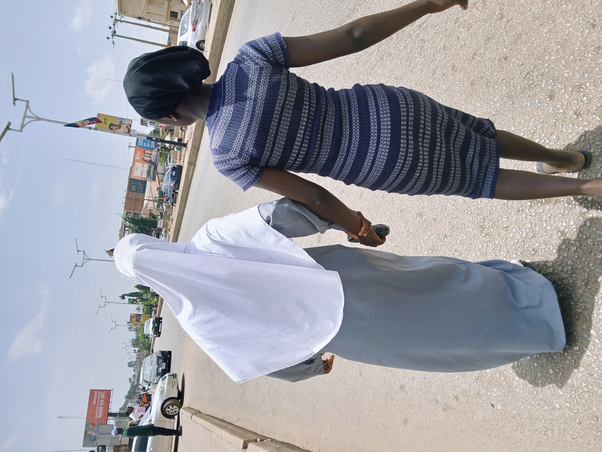 The image depicts two individuals walking on a street in Jos, Nigeria. One person is wearing a blue and white patterned dress and a black head covering, while the other is wearing a white head covering and a grey robe. They appear to be walking side-by-side. The scene includes city infrastructure such as street lights, sidewalks, and roads. Traffic is visible with various cars and billboards with advertising messages, including one for "Detano." Other infrastructure includes buildings and flag poles.