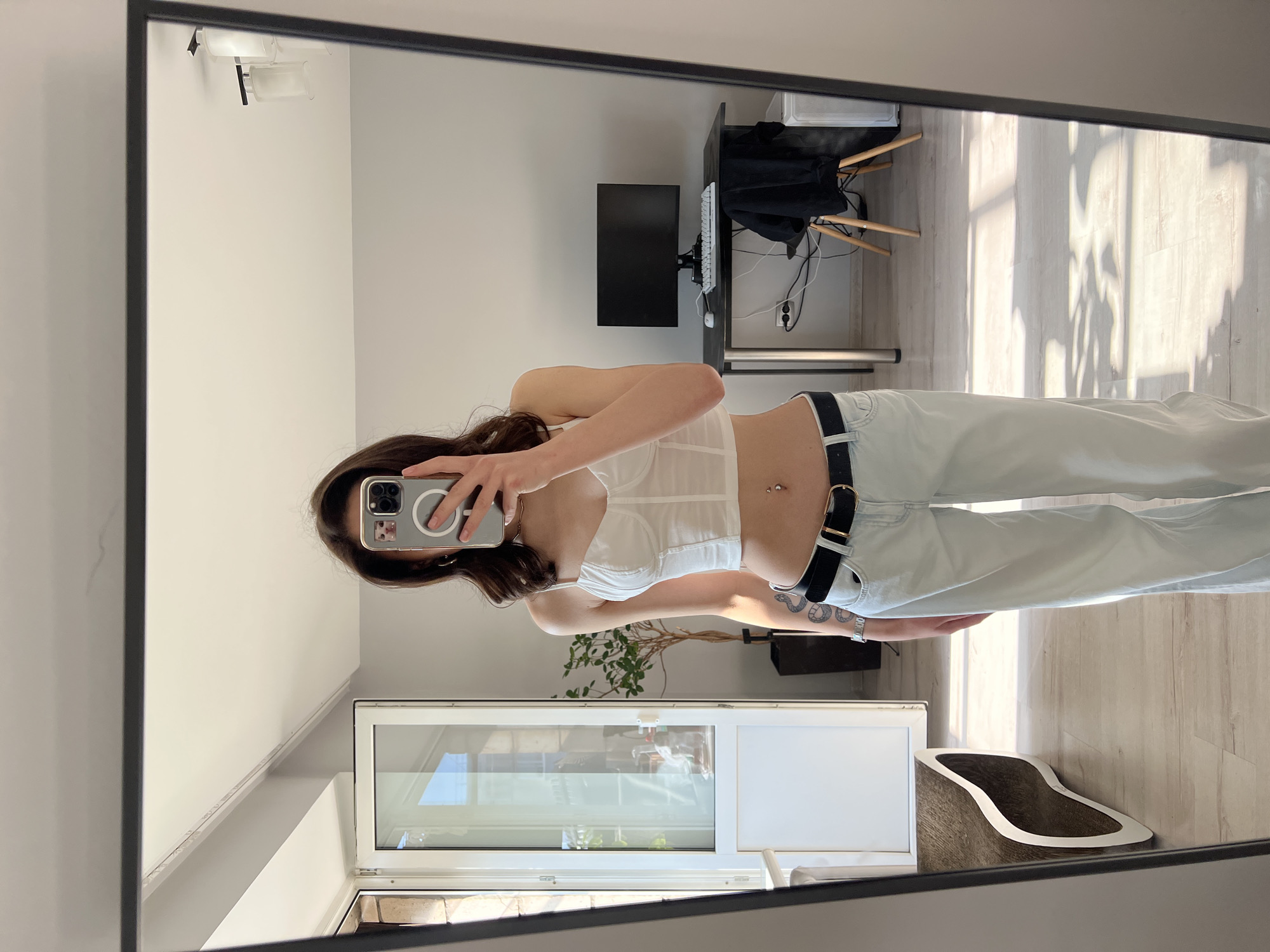 A young woman is reflected in a full-length mirror in a room. She is wearing a white bustier top, a black belt, and light-colored wide-leg jeans. She is holding a smartphone in her right hand, obscuring her face, and taking a selfie. Her left arm has a tattoo of a snake. The room appears to be a bedroom or living area, with light-colored walls and wooden flooring. Sunlight streams in from a window on the left, casting shadows on the floor. In the background, a desk with a computer monitor, keyboard, and mouse is visible, along with a chair and a plant. The room is tidy and appears to be in a residential setting. The image was taken in Ternopil, Ukraine.