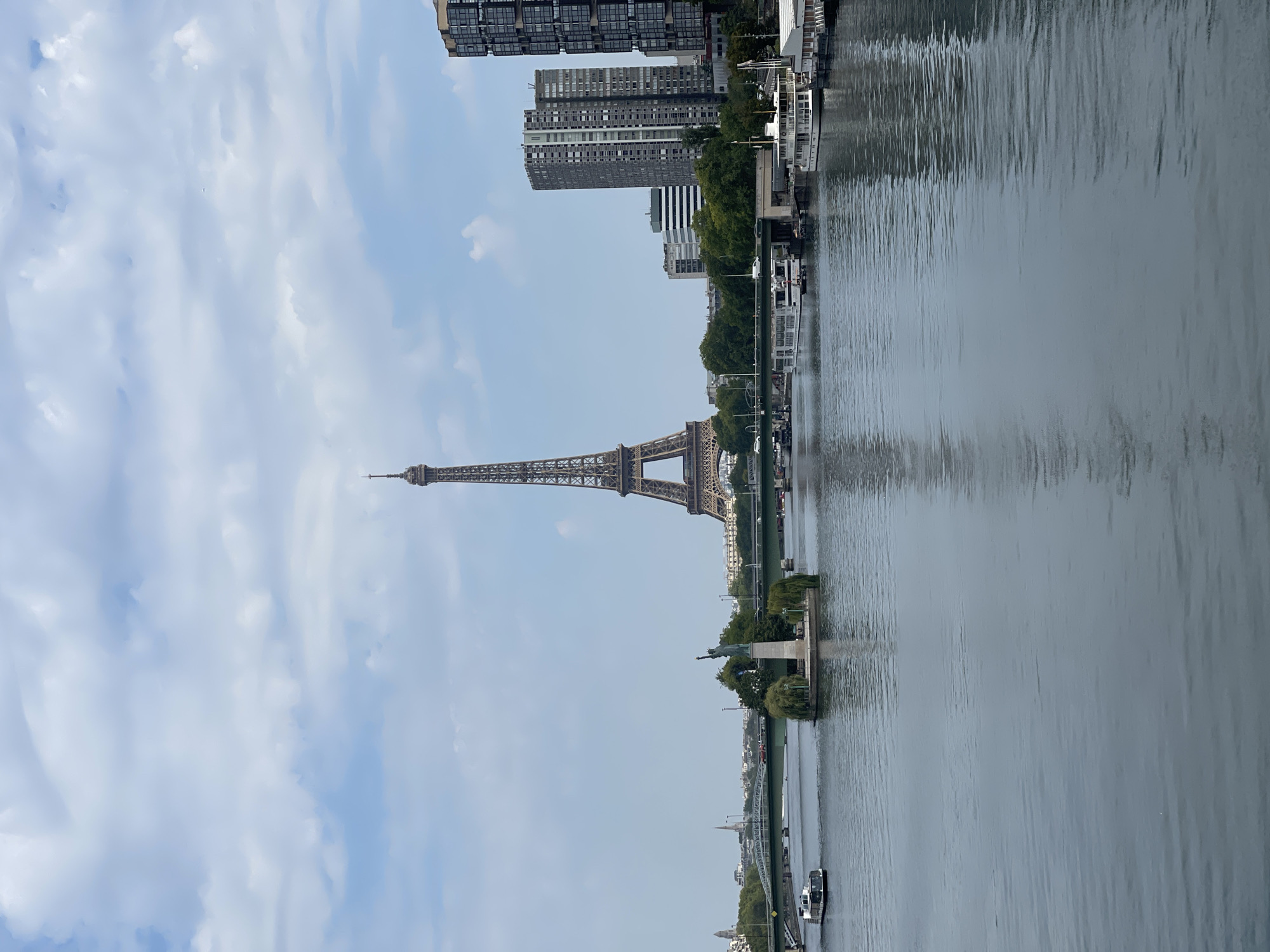 A wide shot shows the Eiffel Tower in Paris, France, situated behind a body of water, likely the Seine River. The sky above is partly cloudy with white cumulus clouds against a pale blue background. The Eiffel Tower, a tall iron lattice structure, is centrally located in the frame, its iconic silhouette dominating the skyline. To the left of the Eiffel Tower, on a small island in the water, stands a replica of the Statue of Liberty, complete with torch and crown.
Several boats are visible on the water. A white tour boat is moving from left to right in the foreground. Further back, other boats are moored along the riverbank, some appearing to be houseboats or docked vessels. A green bridge spans the river in the middle ground, with cars and pedestrians visible crossing it.
On the right side of the frame, modern apartment buildings rise above the trees, their architectural style contrasting with the historic Eiffel Tower. Green trees line the riverbanks, providing a natural element to the urban landscape. The water's surface is calm, reflecting the sky, the Eiffel Tower, and the surrounding buildings, creating a serene and picturesque scene. The overall impression is one of a bustling cityscape with iconic landmarks and daily life unfolding by the river.
