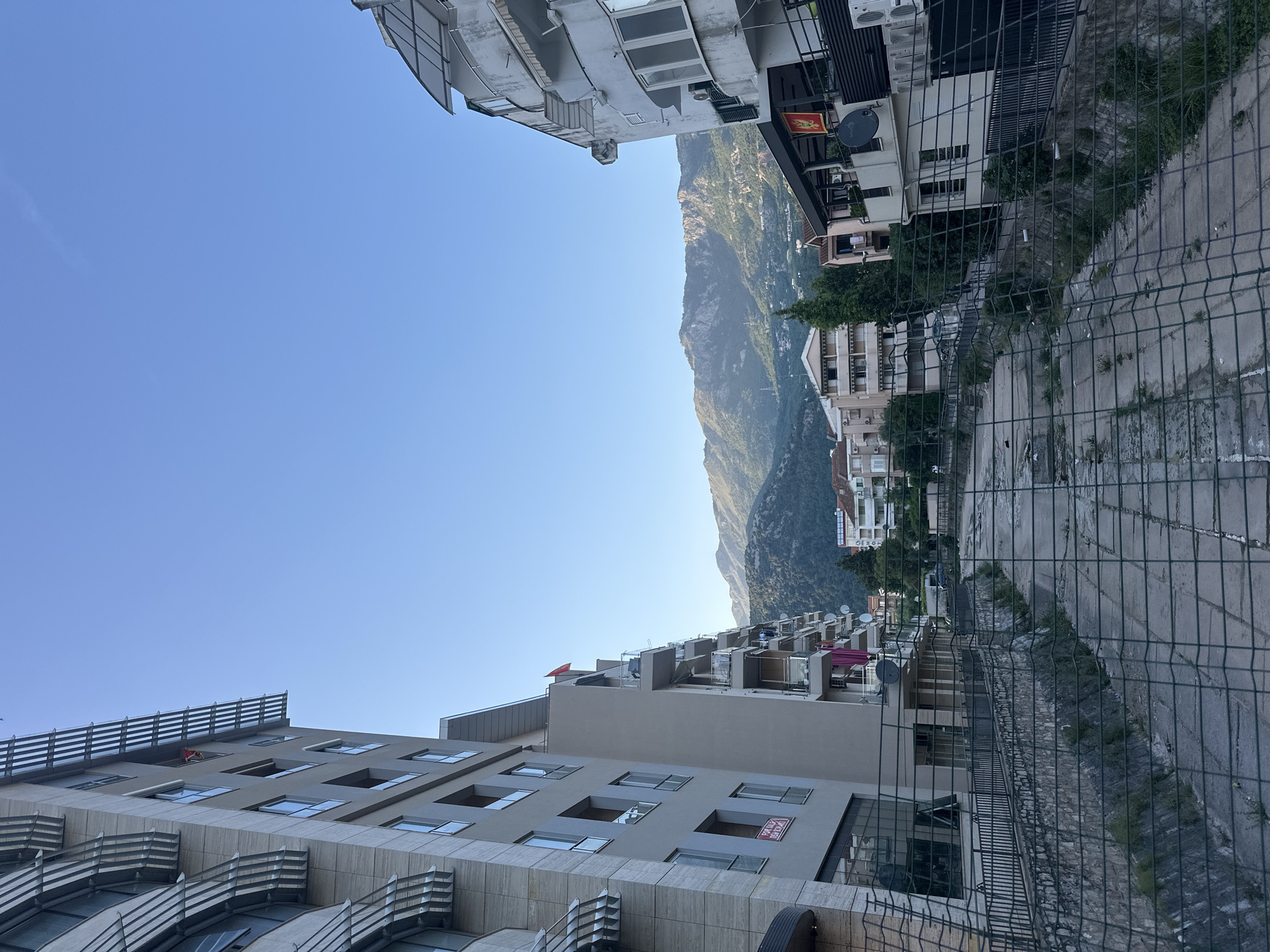 The image depicts a cityscape in Budva, Montenegro, looking towards a mountain range. Buildings flank the view on either side, with the sky visible above. On the left, a multi-story building with a modern architectural design, featuring balconies and large windows, rises into the frame. On the right are older buildings, varying in height and style, partially obscured by a chain-link fence. The mountain range dominates the center of the frame, its peaks rising into the sky. The environment appears to be a mix of residential and urban areas, with a focus on the natural landscape of mountains near built-up spaces. No people are readily visible.