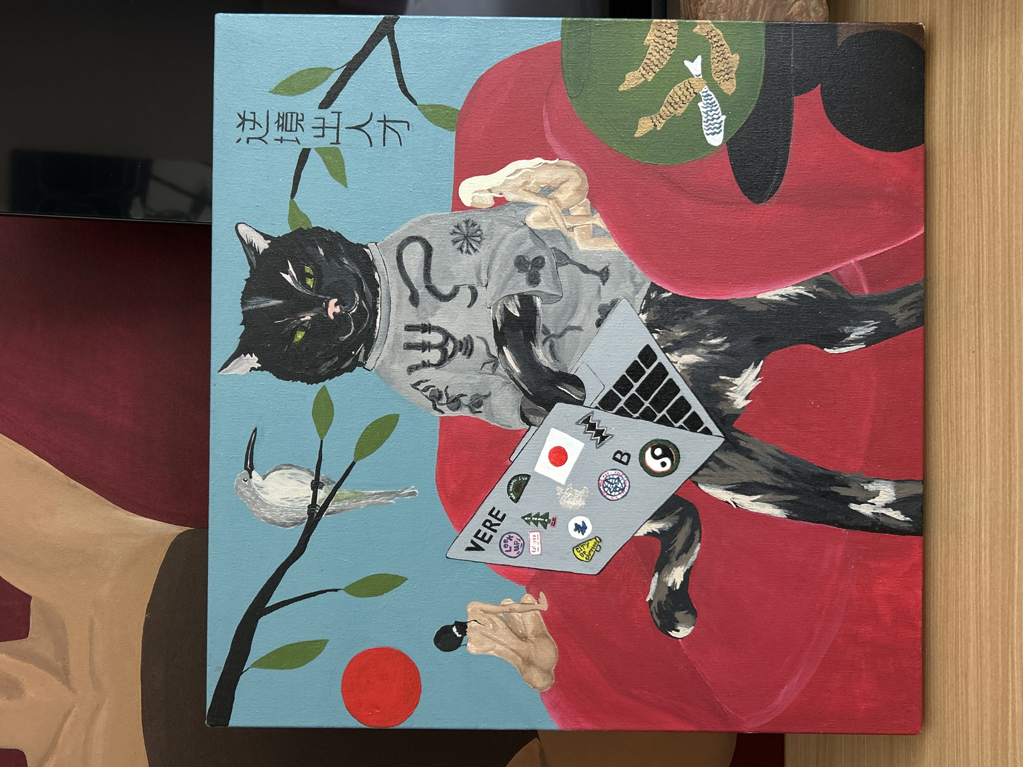 The image is a painting on canvas, depicting a black cat wearing a grey sweatshirt, resting on a red cushion. The cat is lying on its back and holding a laptop on its belly. Stickers are affixed to the laptop, including a Japanese flag, a Yin Yang symbol, and the word "VERE". The cat has green eyes and a pink nose.
The background is split into two sections: the upper left is light blue with tree branches, green leaves, a white bird, and a red circle. In the upper right corner, above the red cushion, is a small green circle with drawings of feet and fish.
The text, "浪裏千丈", is written in vertical lines near the cat's head in the upper left. The image appears to be a painting, and not a photograph of North Kuta, Indonesia. Therefore, no details about North Kuta, Indonesia can be provided.