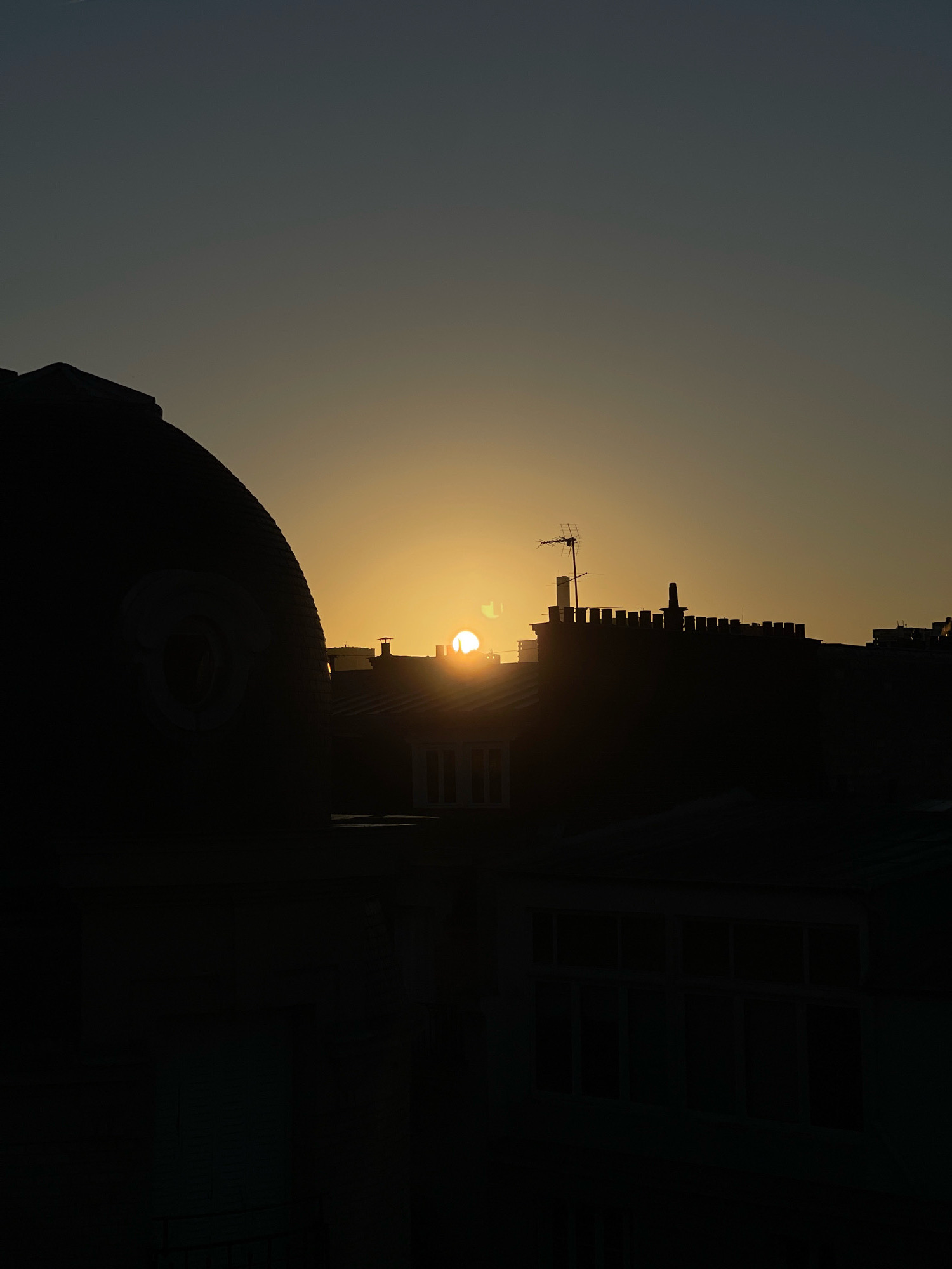 The image captures a silhouette of Parisian rooftops against a twilight sky. The setting sun is partially visible, casting a warm, golden glow that illuminates the upper atmosphere. In the foreground, the dark, curved silhouette of a dome, possibly part of a historic building, dominates the left side of the frame. To the right, a series of rooftops with crenellated parapets and chimneys are silhouetted against the sky. An antenna stands prominently on one of the buildings. The lower portion of the image shows the dark outlines of more buildings, including windows that appear lit from within or reflect the fading light. The overall scene depicts an urban landscape at dusk, with the fading sunlight creating a dramatic and serene atmosphere.