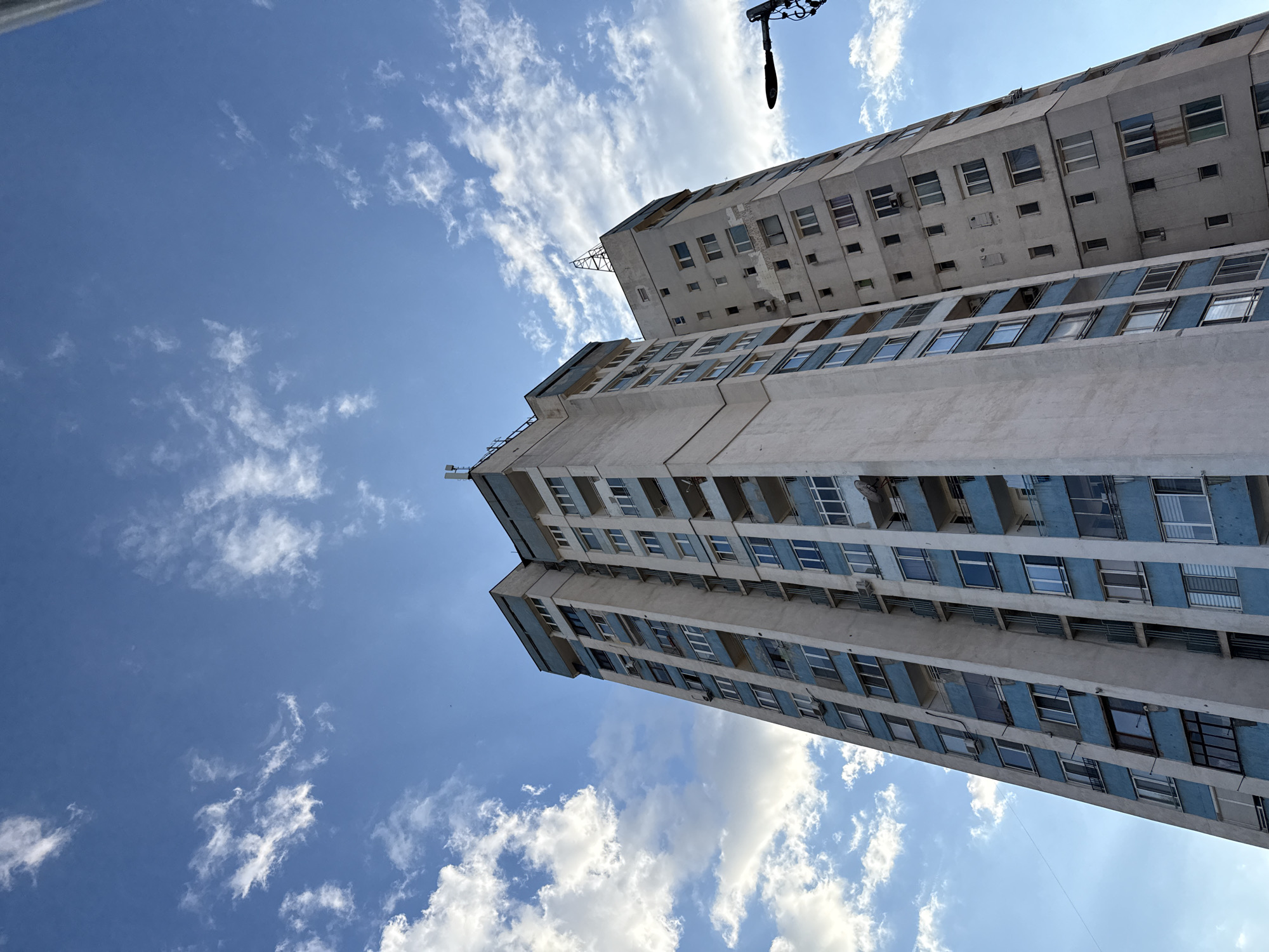 The image shows a tall, multi-story building complex in Sofia, Bulgaria, angled against a blue sky with scattered white clouds. The building's facade is predominantly white or light gray, with rows of windows, some of which have blue accents around them. The building appears to be constructed in a Brutalist architectural style with a somewhat worn or aged appearance. There is a dark, possibly metal object in the top right corner of the image, slightly obscuring the view of the building. The perspective suggests the photo was taken from a low angle, looking upwards. There are no people visible in the frame.