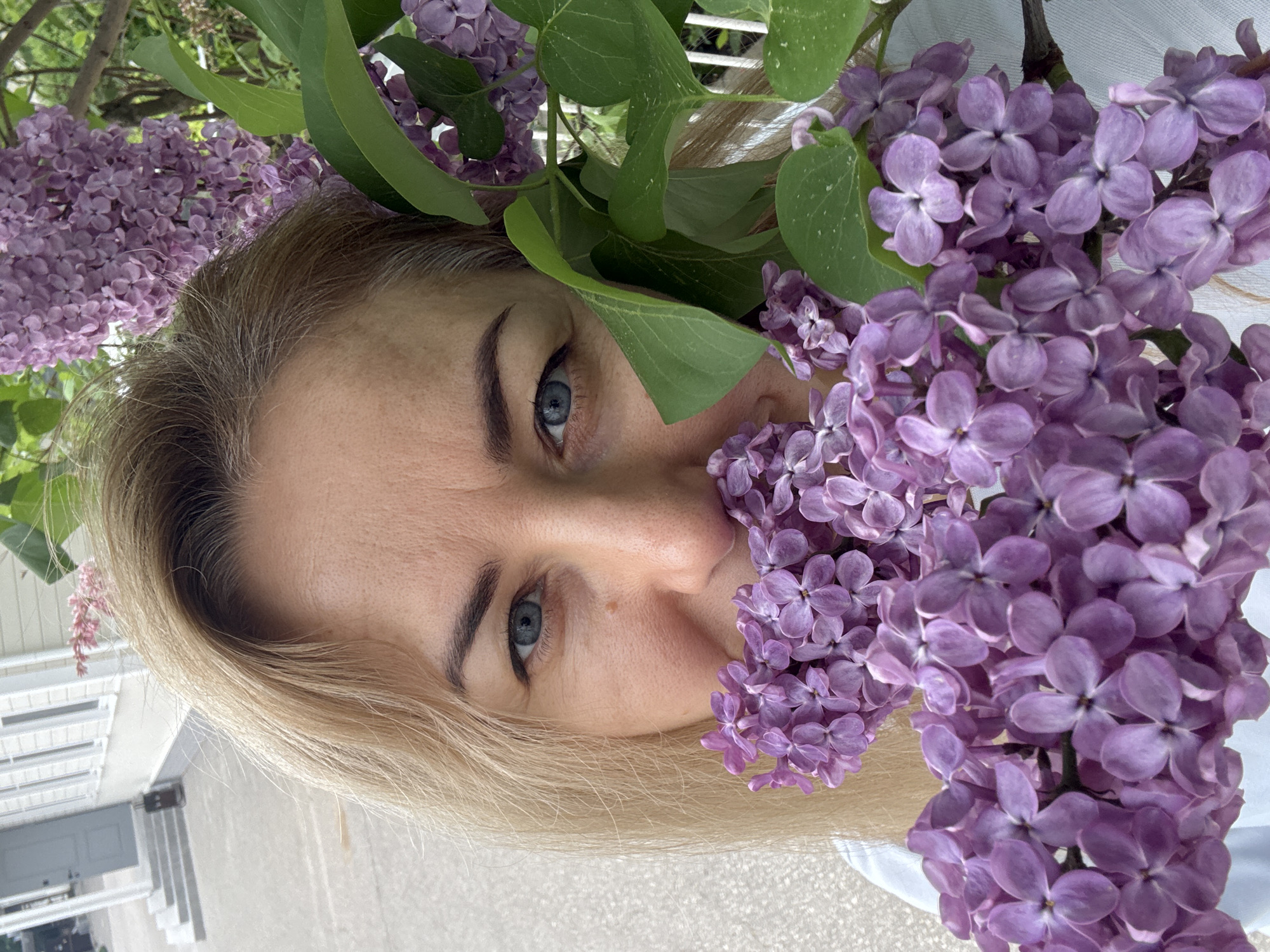 A woman with blonde hair and blue eyes poses with lilac flowers. The scene appears to be in a garden setting. The woman's face is partially obscured by the purple flowers. In the background, a house with white walls and visible steps can be observed. The location is inferred to be in the vicinity of Боголюбово, Russia.