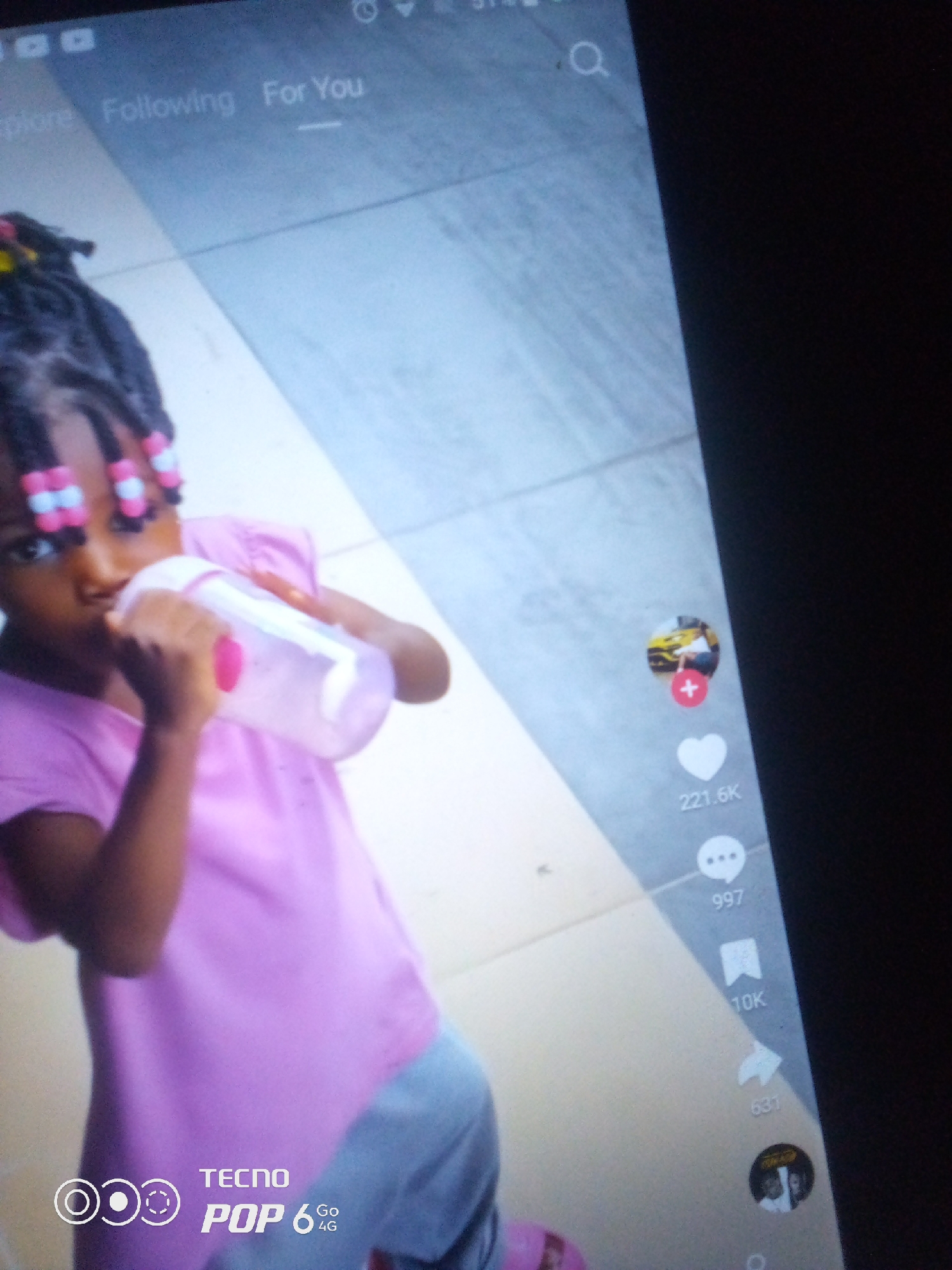 The image shows a young child, likely female, with braided hair adorned with pink and white beads, drinking from a pink and white bottle. The child is wearing a light pink shirt and light blue pants. The background appears to be an indoor setting with a tiled floor. On the right side of the image, social media icons are visible, including a profile picture, a play button with a car, a heart with "221.6K" underneath, a comment icon with "997" below it, a bookmark icon with "10K" underneath, and a share icon with "631" next to it. At the bottom left, a "TECNO POP 6 Go 4G" logo is present.
Based on the provided information, it is impossible to determine the location within the city of Howayi, Nigeria. The image itself does not contain any visual cues that would allow for such a specific localization.