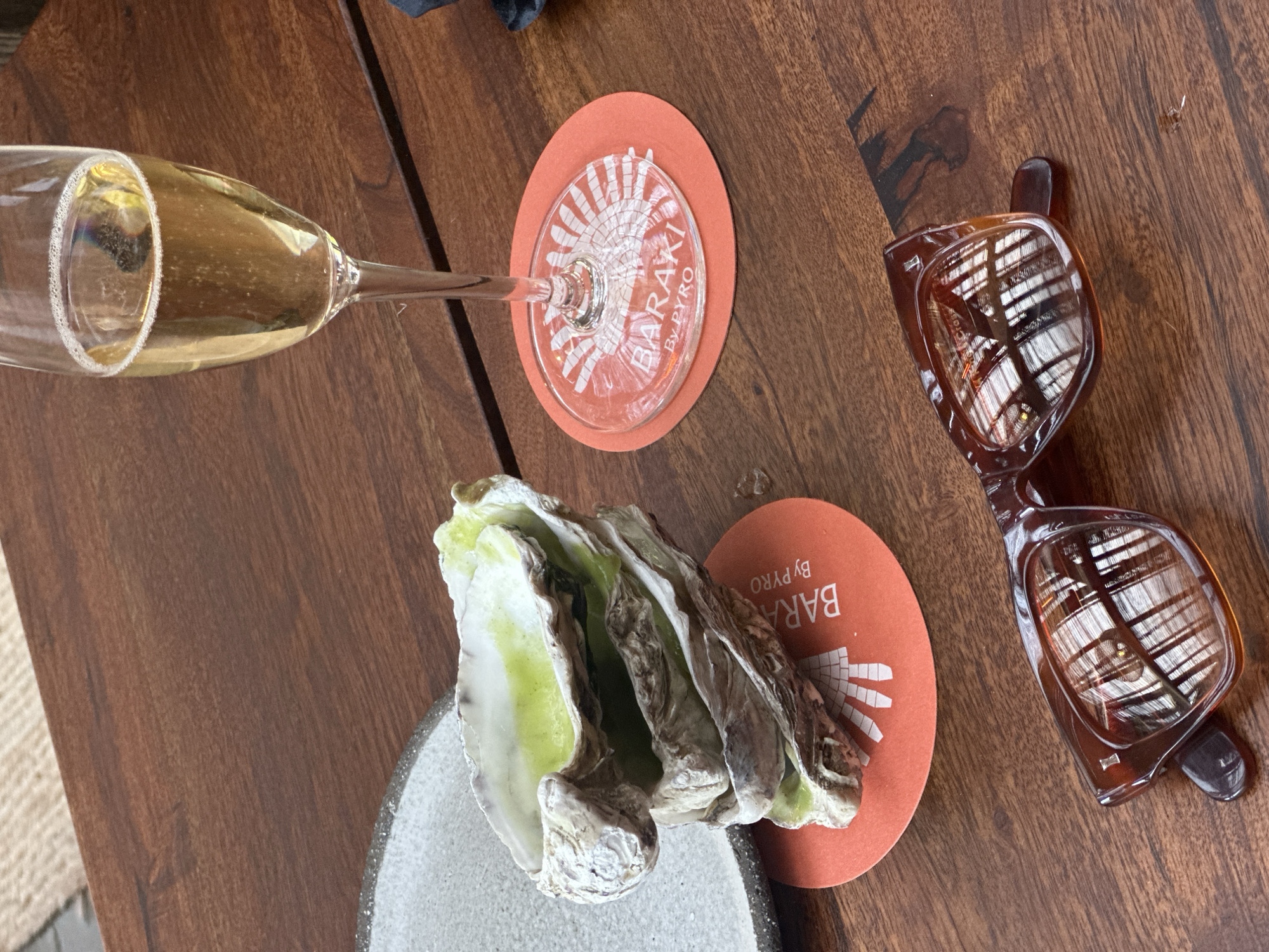 The image depicts a table setting with a glass of champagne, an oyster on a plate, coasters with the "BARAKI By PYRO" logo, and a pair of tortoiseshell sunglasses. The setting suggests a restaurant or bar in London, United Kingdom, likely affiliated with "BARAKI By PYRO." The objects are arranged on a wooden table.