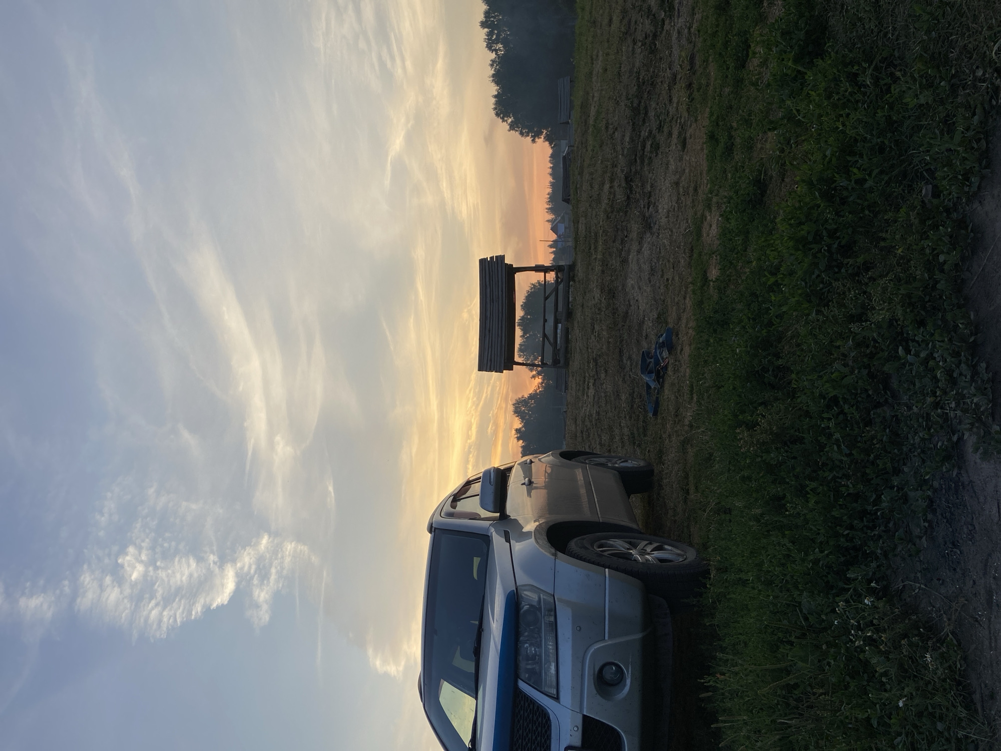 In Peshcherka, Russia, a silver SUV is parked on a grassy field under a sky with wispy clouds and the soft glow of sunset. In the background, a wooden gazebo with a weathered roof stands on a slight incline, with a row of trees forming a dark silhouette against the orange and yellow horizon. Two pairs of dark sandals lie discarded on the dry grass near the gazebo. The foreground is dominated by lush green grass, with the lower portion of the SUV visible on the left. The scene depicts a tranquil, rural outdoor setting during twilight.