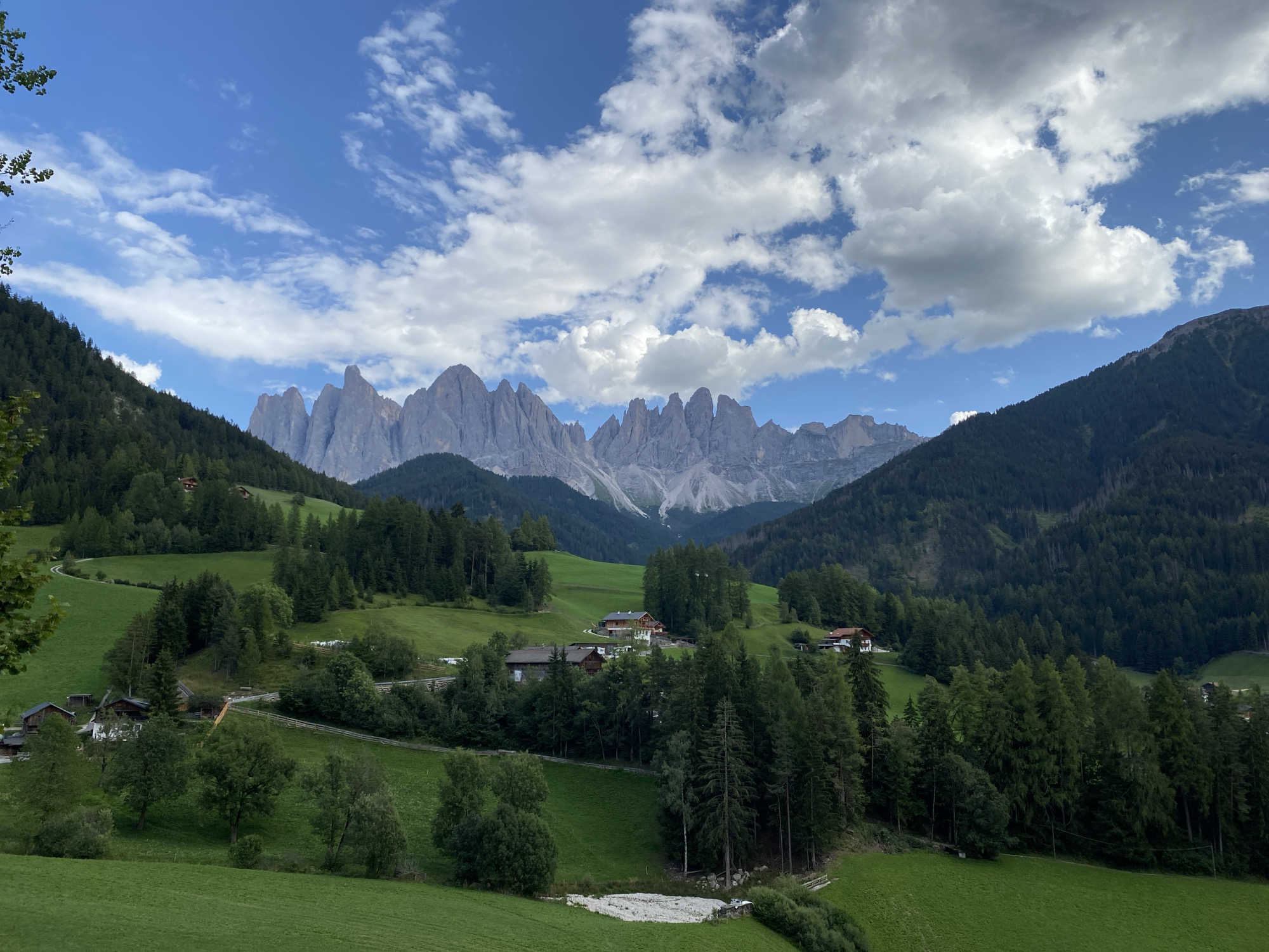The image depicts a picturesque landscape of the village of St. Magdalena (Santa Maddalena) in Italy. The scene is dominated by rolling green hills dotted with scattered houses and dense pine forests. In the background, a dramatic mountain range with jagged peaks rises against a bright blue sky filled with fluffy white clouds. The foreground features well-maintained green meadows and a winding dirt path. The overall impression is one of serene natural beauty and rural charm.
