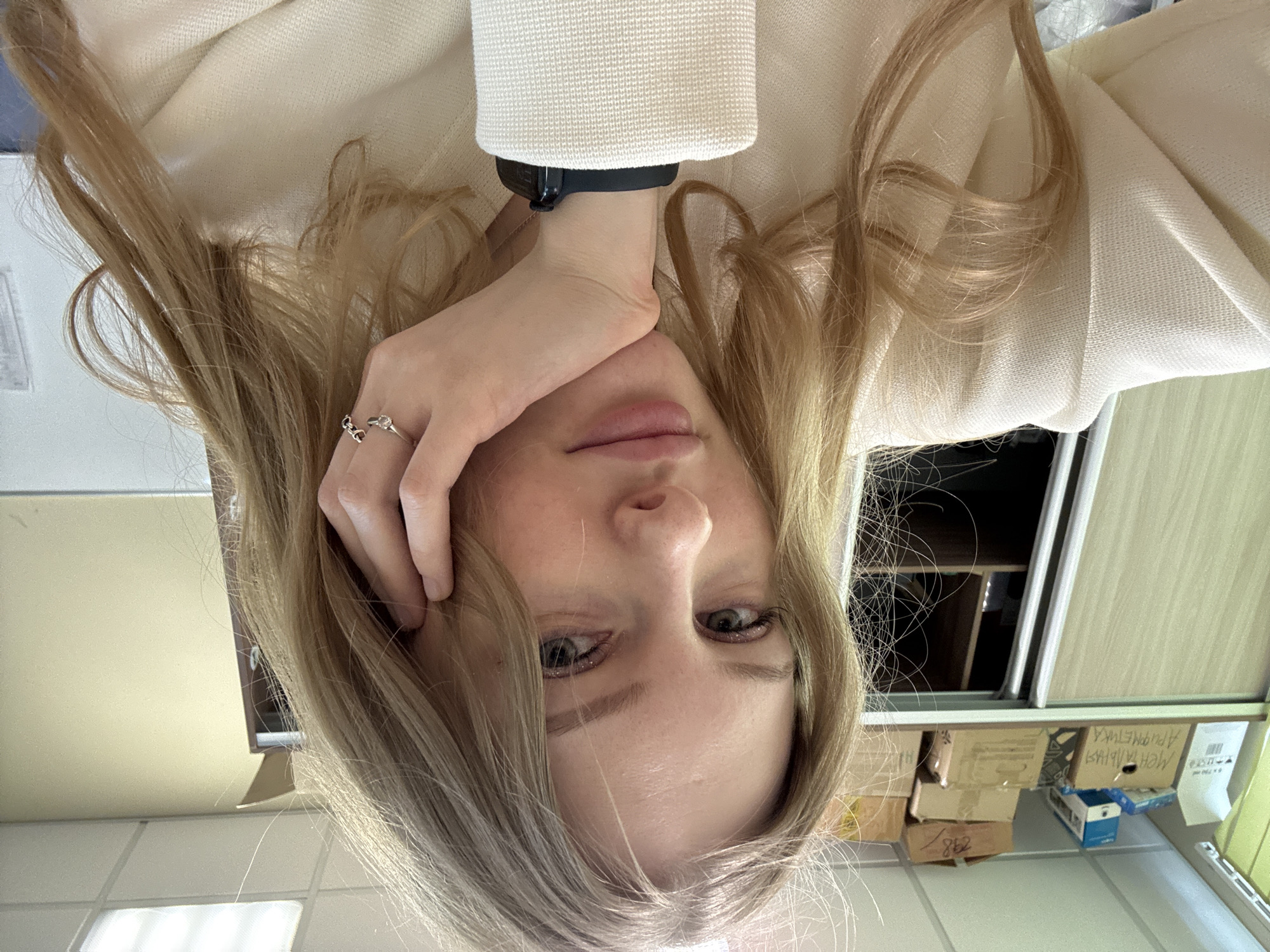 A young woman with long blonde hair is posing for a selfie. She has fair skin and is wearing a ring on her left hand. She is wearing a white jacket and a black smartwatch on her left wrist. The background appears to be an office or work space with beige and white walls, boxes, and storage cabinets. The location is potentially Perm, Russia, but this is only based on the image content and cannot be definitively confirmed without additional information.