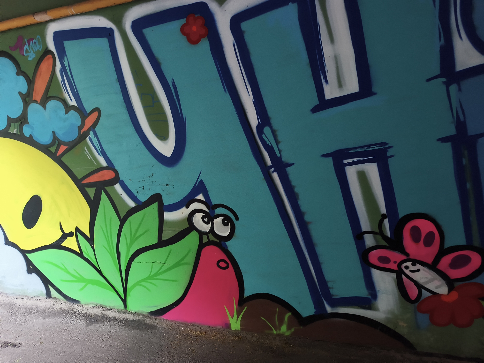 Graffiti art adorns a wall, likely in Kyiv, Ukraine. Dominating the view is a large, stylized blue letter "H" outlined in navy, with white highlights. Adjacent to the "H" is a cartoonish scene featuring a yellow shape with a black eye, surrounded by green leaves. A pink, snail-like figure with large googly eyes is also part of the composition. A butterfly with pink and dark red wings, as well as a small red flower, is drawn near the letter. The background is a mix of teal and green. The bottom of the wall transitions to a darker gray surface, suggesting a ground level or sidewalk.