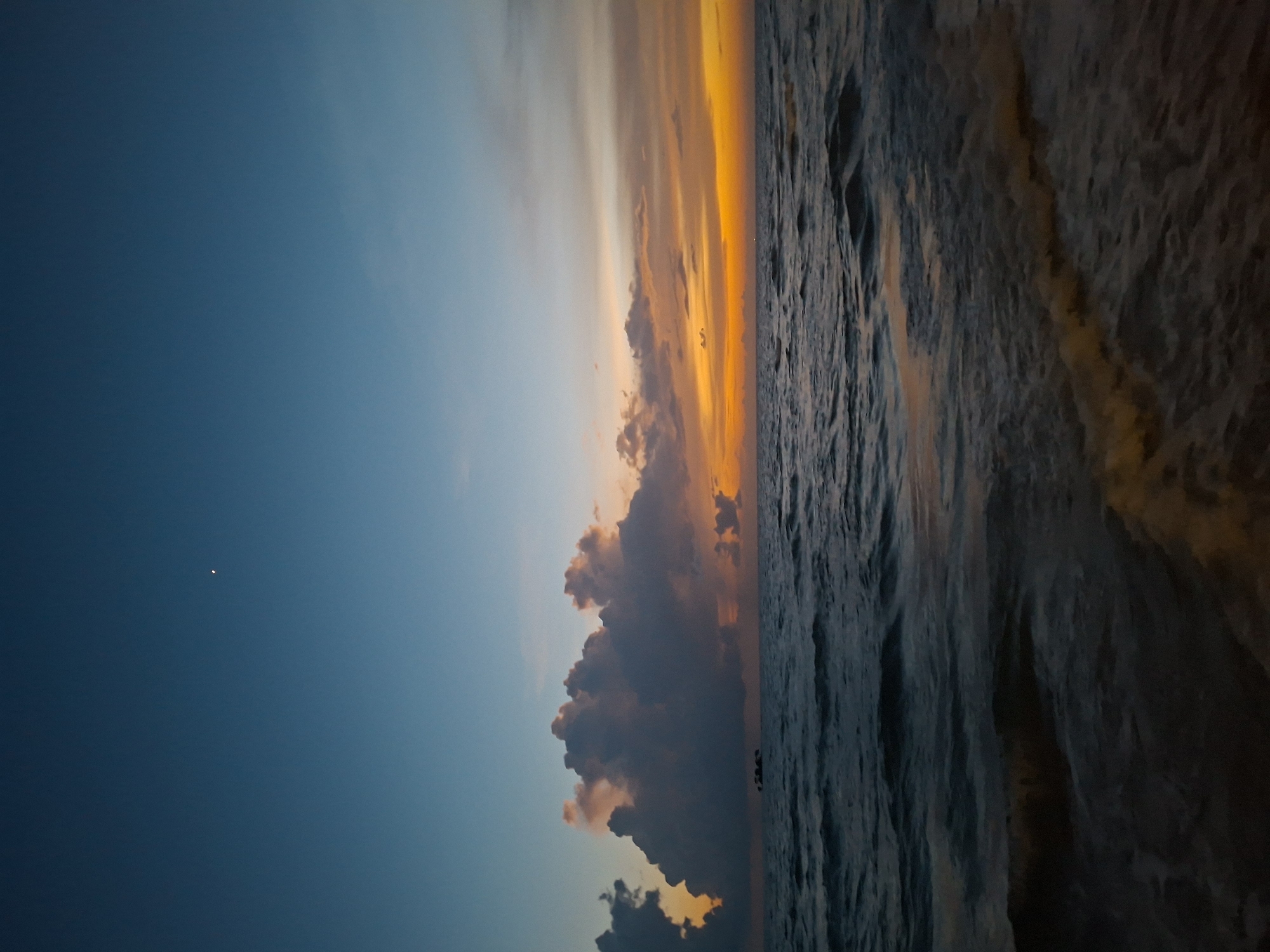 The image depicts a serene seascape at dusk. The sky transitions from a deep blue at the top to a vibrant orange and yellow horizon, indicating the setting sun. Large, dramatic clouds are scattered across the sky, with some illuminated by the sunset's glow. A small, distant object, possibly a boat, is visible on the water near the clouds. The ocean surface is textured with gentle waves, reflecting the twilight colors. The foreground shows the edge of a beach or shallow water with subtle wave patterns. The overall atmosphere is tranquil and picturesque. The location is not specified within the city of Unknown, Unknown.