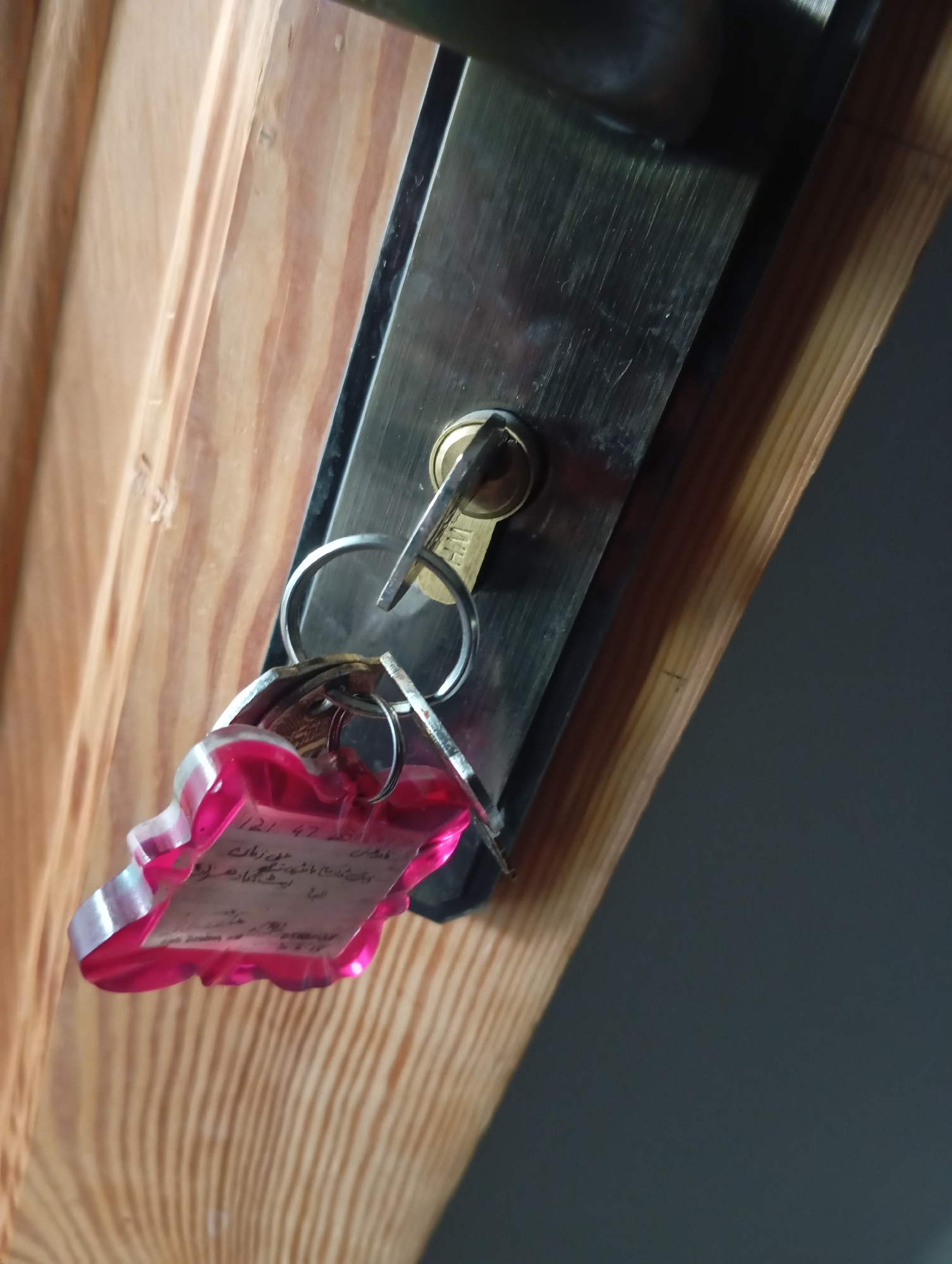 A close-up, angled shot shows a set of keys hanging from a metal door lock mechanism. The lock is mounted on a light-colored wooden door with visible grain. The keys are attached to a pink, heart-shaped tag with writing on it. One of the keys is partially inserted into the lock. The background is a dark, out-of-focus area. The scene appears to be indoors, likely in a residential or commercial building, as indicated by the door and lock. The lighting is dim, suggesting it could be nighttime or an interior space with minimal natural light.