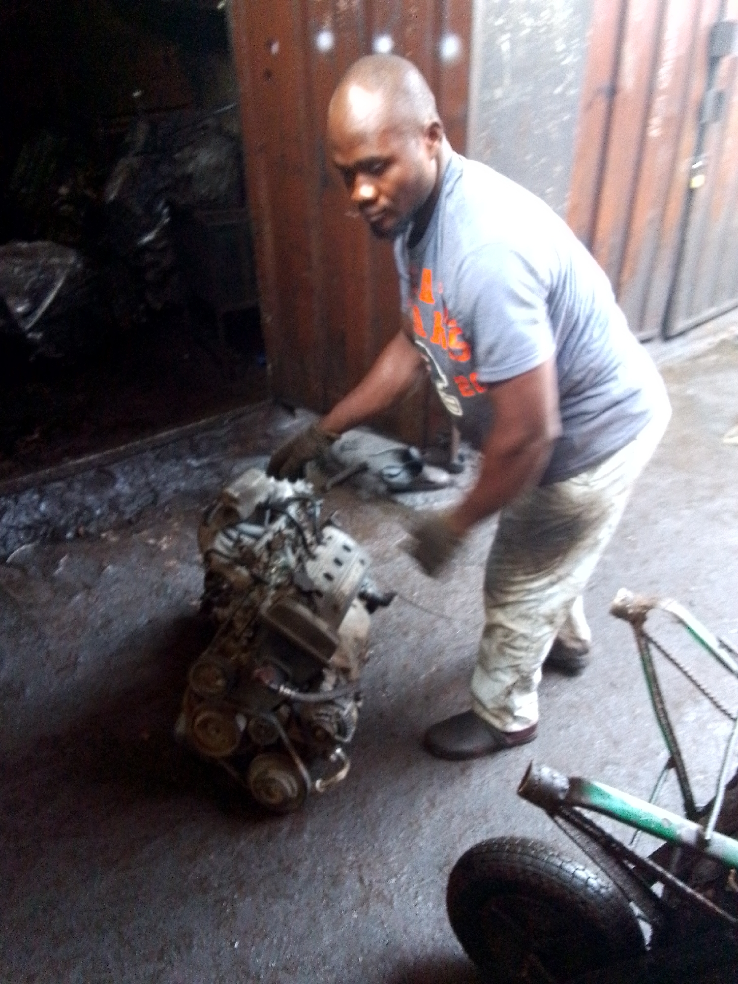The scene takes place outdoors, presumably in the city of Onitsha, Nigeria, indicated by the presence of the male individual in the image. The main subject is a man, wearing a gray t-shirt with orange lettering and camouflage-patterned pants. He's bending over, with his gloved hands near a greasy car engine on the ground. The ground appears to be concrete. Next to him, partially in the frame, is a green hand truck or dolly, with a tire visible. Behind the man is a dark space, potentially an open garage or storage area, with miscellaneous items inside. A corrugated metal wall is also visible behind the man.