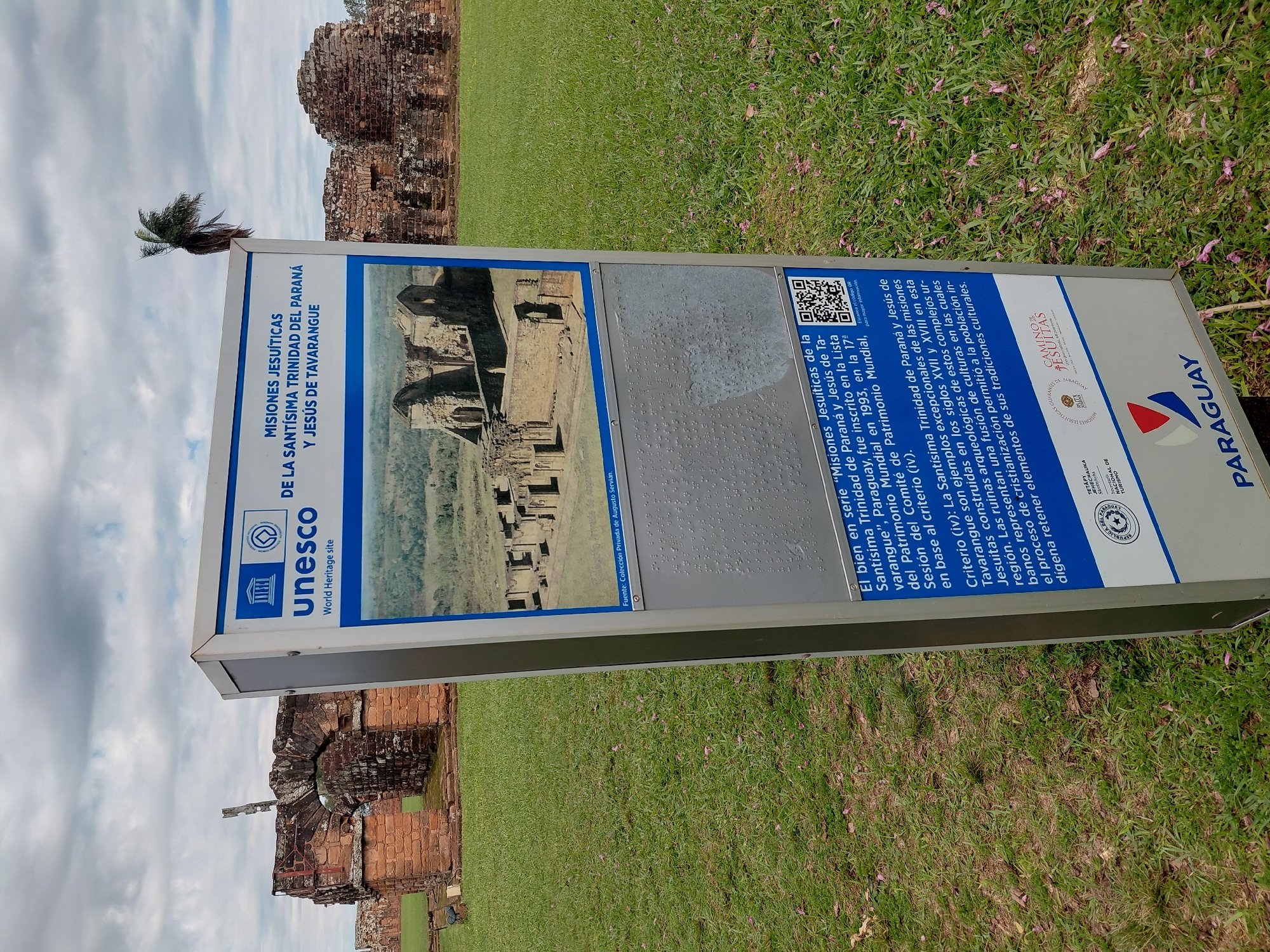 The image shows a weathered information kiosk in front of the ruins of a Jesuit mission in Trinidad, Paraguay. The kiosk has multiple panels with text and images, including a UNESCO logo and a photograph of the ruins. The background shows green grass, scattered pink flower petals, and more crumbling brick structures of the mission. The sky is cloudy. The kiosk is made of metal and plastic, with some panels showing signs of wear and tear. The text on the kiosk is in Spanish and appears to be about the history and significance of the mission. A QR code is visible on one of the panels. The ruins themselves are made of red brick and are partially overgrown with vegetation. A lone palm tree is visible in the distance. The scene suggests a historical site open to public visitation.