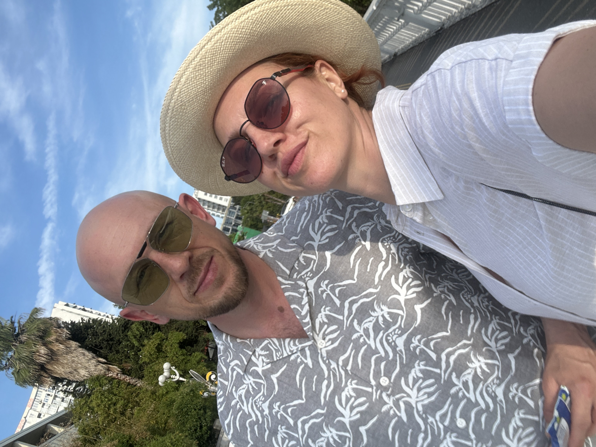 The image depicts a selfie of a man and a woman taken outdoors on a sunny day in Sochi, Russia. The man, bald with light stubble, wears aviator-style sunglasses with yellow-tinted lenses and a grey short-sleeved shirt patterned with white palm trees and waves. He is positioned slightly behind and to the left of the woman, with his left hand holding a blue and white patterned object at the bottom left of the frame. The woman wears a wide-brimmed straw hat, round sunglasses with reddish lenses (a "Ray-Ban" logo is visible on the left lens), and a light-colored, vertically striped short-sleeved button-up shirt. She has reddish hair visible and a pearl earring in her right ear, with her lips slightly puckered in a playful expression.
In the background, under a clear blue sky with sparse white clouds, are lush green trees, including a tall palm tree with a shaggy trunk. Behind the foliage, multi-story white residential or hotel buildings are visible. To the left, a decorative street lamp with three globe lights and a yellow-and-black bee-like sculpture is present. The subjects are standing on a walkway or bridge, indicated by a white painted railing visible along the right side of the image.