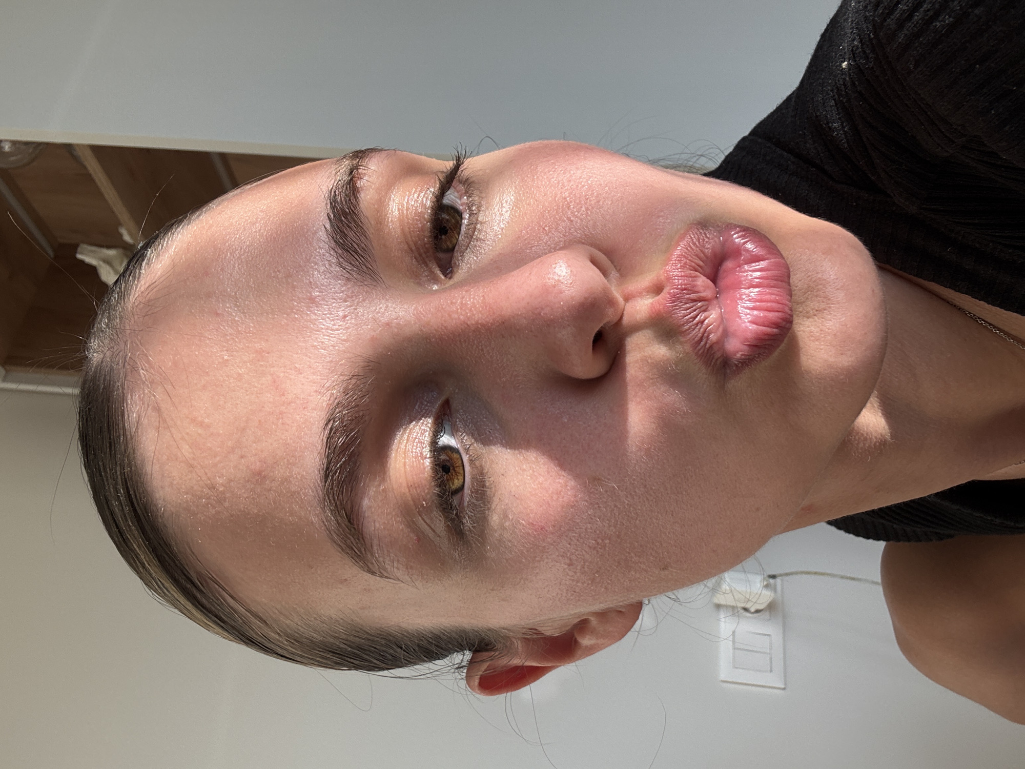 A close-up, angled portrait shows a young woman indoors in Unknown, Unknown. Her head is tilted significantly to the right. She has fair skin with a slightly oily sheen, defined eyebrows, and brown eyes. Her lips are puckered in a kissing gesture. She is wearing a black, ribbed top. The background shows a white wall with an electrical outlet. The upper left corner shows a partial view of a ceiling. The image is well-lit with direct sunlight.