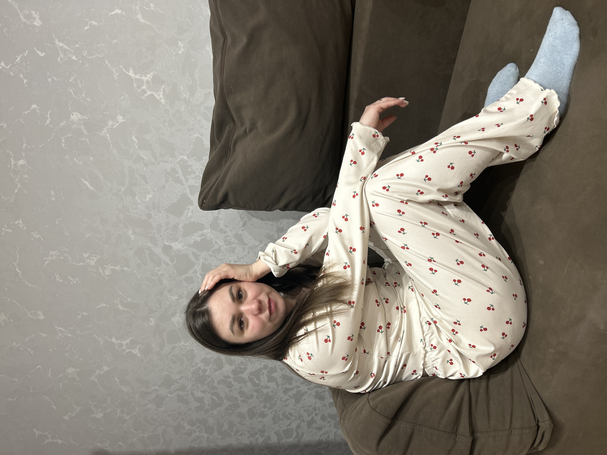 A woman is reclining on a brown couch. She is wearing cream-colored pajamas with a cherry pattern and light blue socks. She has brown hair and is touching her head with her hand. A brown pillow is beside her on the couch. The background is a gray wall with a marbled pattern. The scene is indoors. The location may be Chaikovsky, Russia.