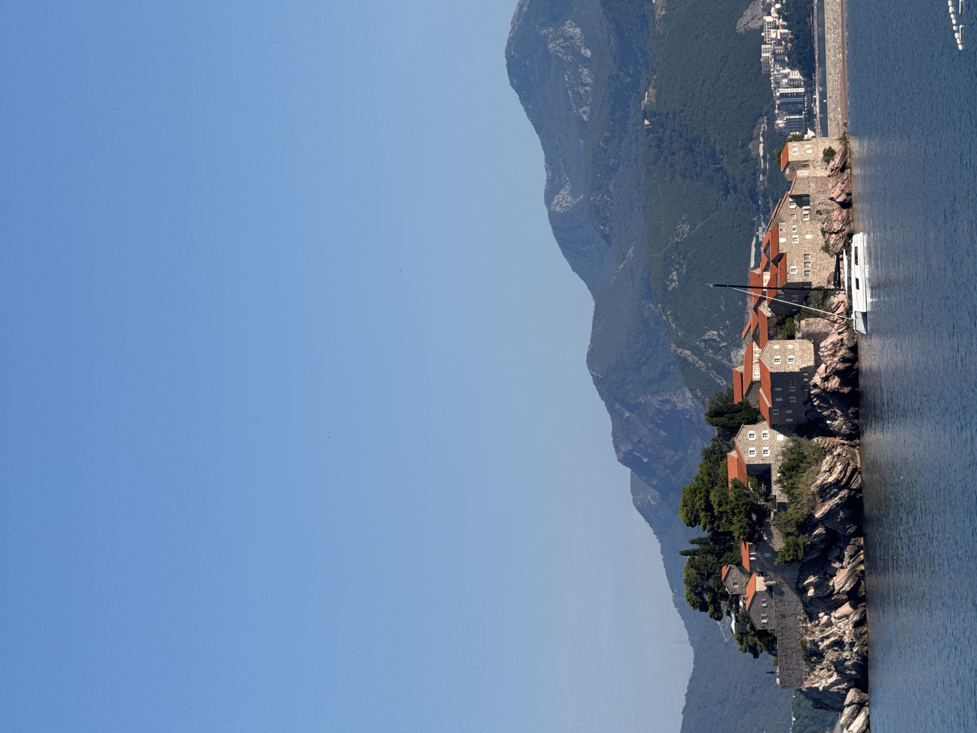 The image shows Sveti Stefan, a fortified island village in Montenegro, situated on a rocky outcrop connected to the mainland by a narrow causeway. The island is densely packed with stone buildings featuring distinctive red-tiled roofs. Lush green trees and vegetation sprout from the rocky terrain of the island. In the foreground, calm blue water laps against the rocky base of the island. A white catamaran sailboat is moored to the side of the island. In the background, a town with modern buildings is visible along the coastline, and beyond that, rolling green hills and mountains rise under a clear blue sky. The scene captures a sunny day with excellent visibility.