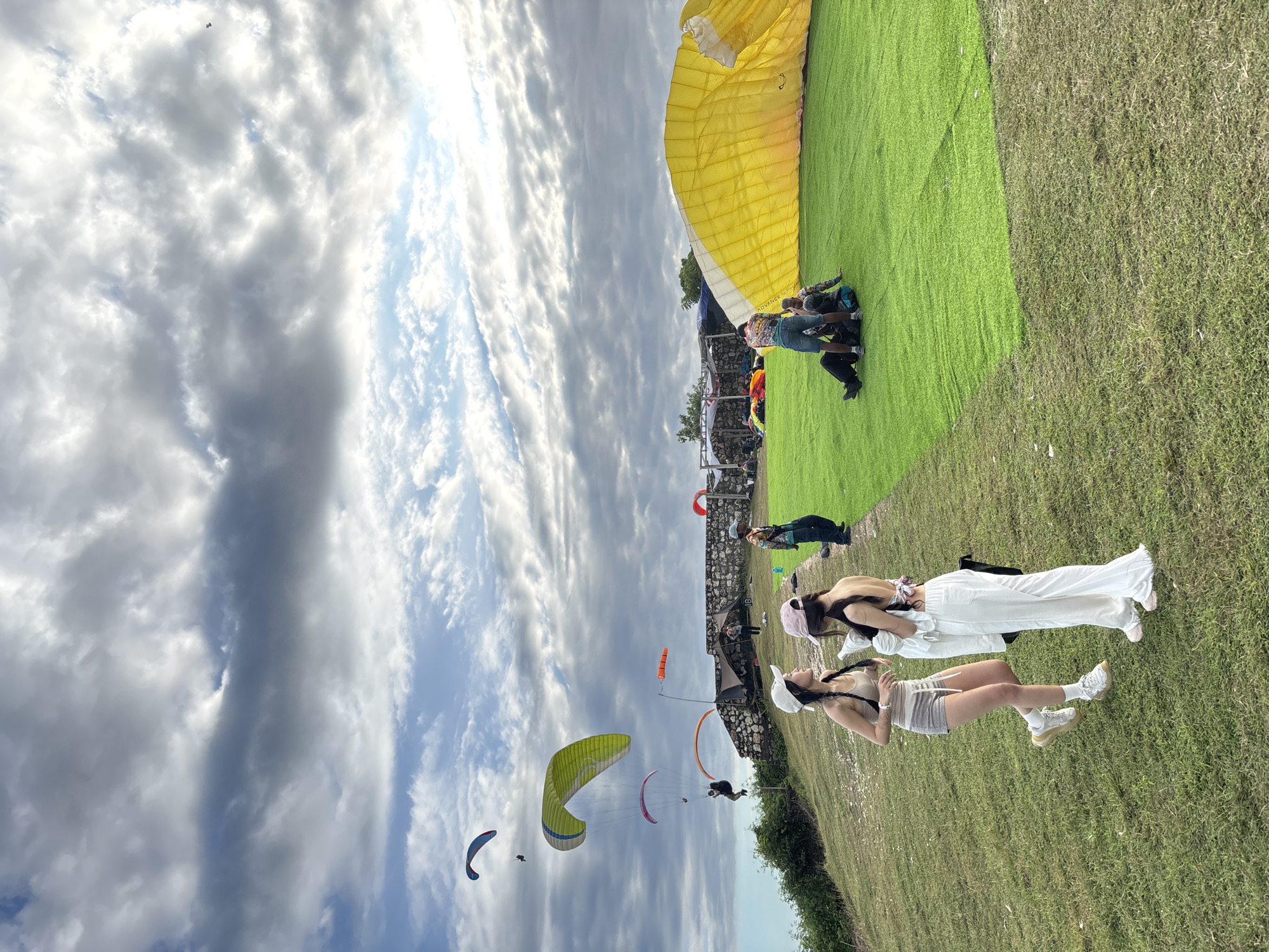 Two women are posing on the grass in Kuta Selatan, Indonesia, near paragliding activities. The women are wearing casual clothing and hats. Several paragliders are in the sky. A yellow paragliding wing is on the grass in the background. Other people are present near the wing. The sky is cloudy.