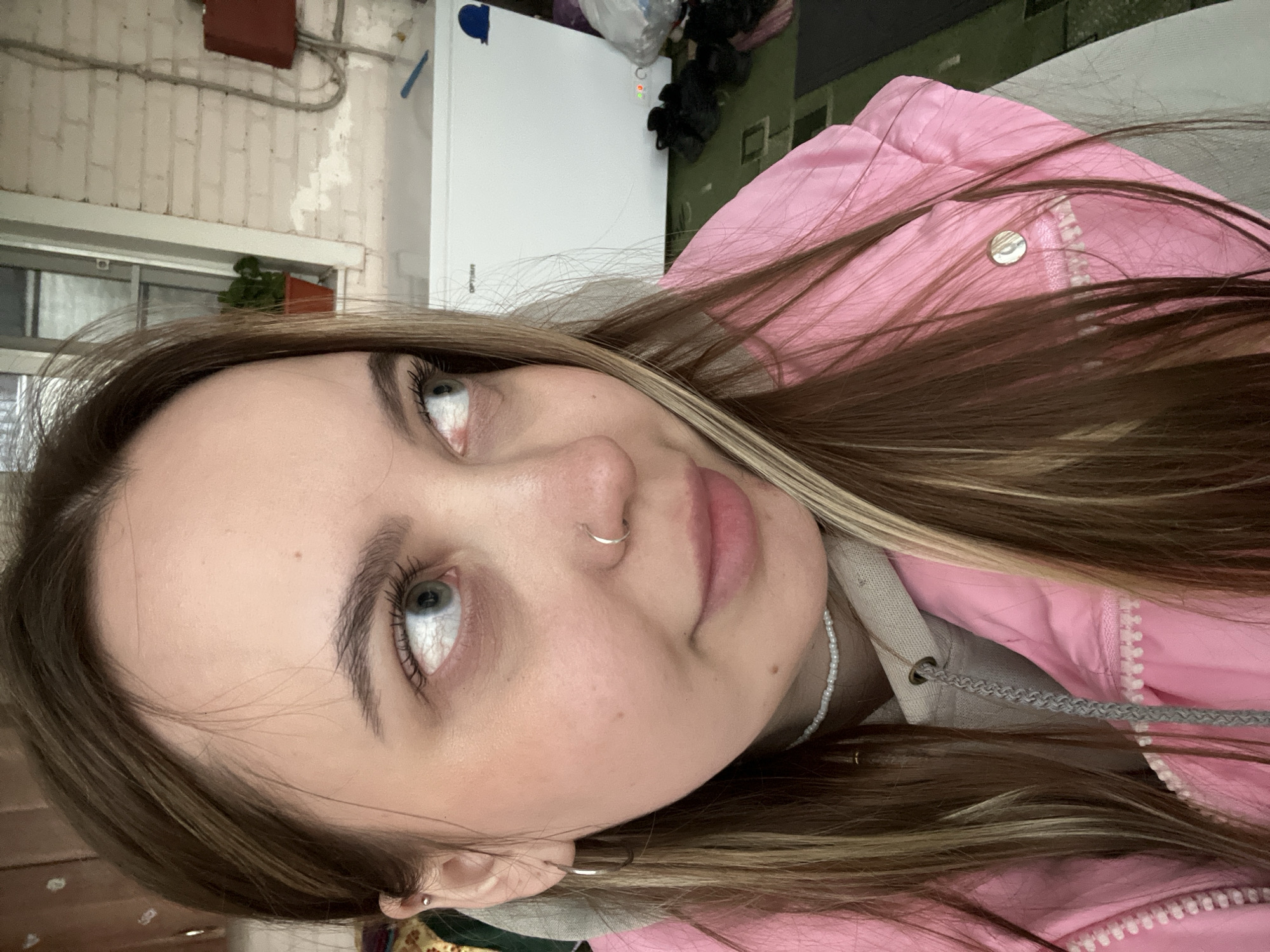A young woman is photographed indoors. She has fair skin, light brown hair with blonde highlights, and light-colored eyes. She has a ring in her nose and earrings in her ears. She is wearing a pink jacket over a gray hooded sweatshirt, and a white beaded necklace. The background includes a white refrigerator, a brick wall, a window with plants, and dark-colored flooring. The location is not evident from the image to be Novopokrovskaya, Russia.