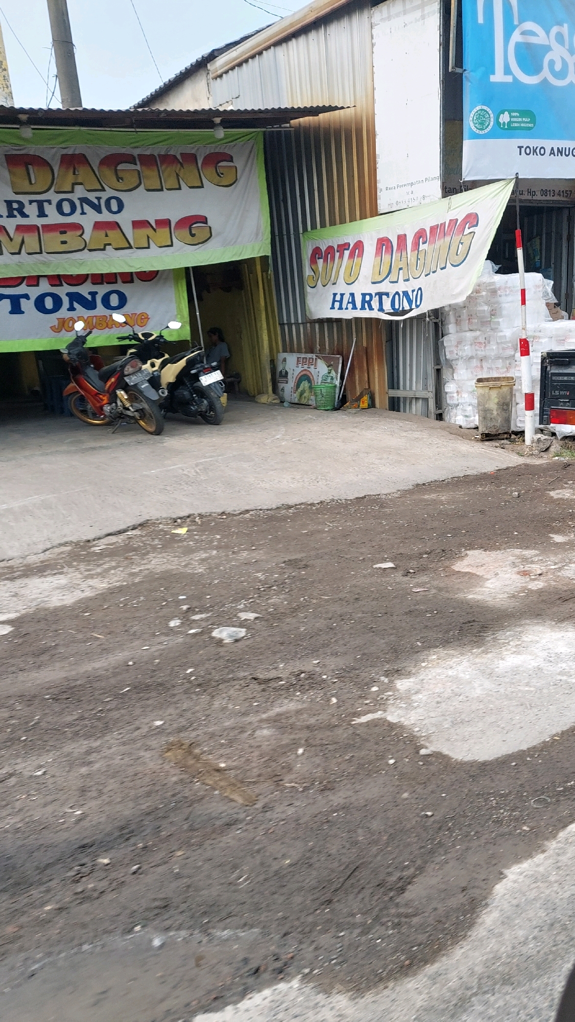 The image shows a street scene in Pilang, Indonesia, in front of a food establishment. A large banner with green and yellow lettering hangs above the entrance, advertising "SOTO DAGING HARTONO." Another banner on the right side of the building also displays "SOTO DAGING HARTONO." Two motorcycles, one orange and one beige, are parked in front of the entrance. A person is visible inside the establishment. Stacks of white styrofoam containers are piled to the right of the building, along with a red and white striped pole and a brown trash bin. In the background, a white building with blue lettering that reads "Tess" is visible. The ground in the foreground is a rough, unpaved surface with patches of dry earth and puddles of water.