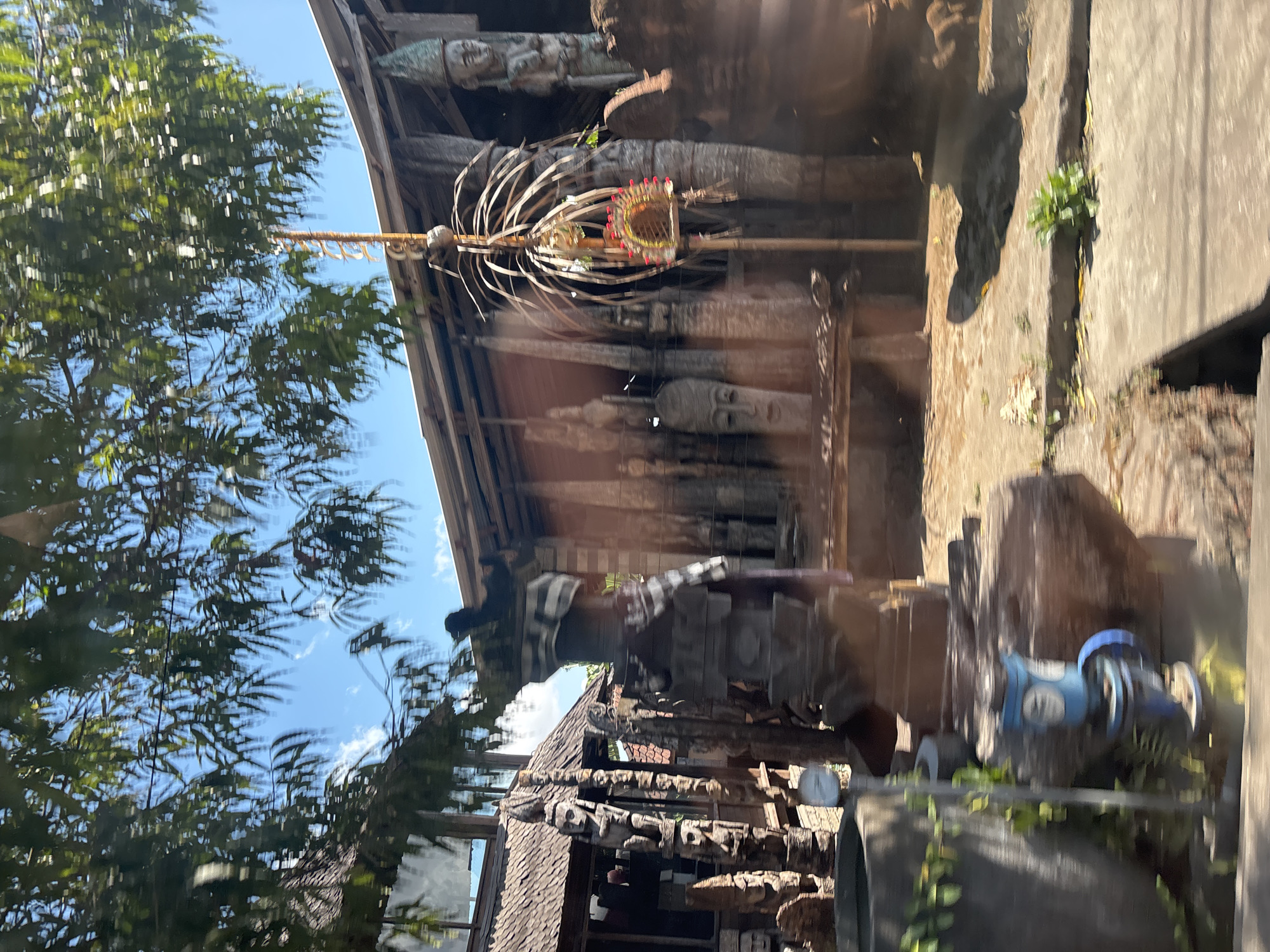 The image shows a street scene in Sukawati, Indonesia, featuring an outdoor display of wooden sculptures and traditional Balinese architecture. The sculptures, predominantly carved from dark wood, include figures with distinct facial features, some appearing to be totemic or ancestral representations. They are arranged in front of a building with a thatched roof and wooden walls. A tall, slender bamboo-like structure with a decorative element at its top stands centrally, adorned with red and yellow accents. Green foliage from overhanging trees partially obscures the sky, which is blue with scattered white clouds. In the foreground, a large, weathered stone pot and a blue metallic object with a gauge are visible, suggesting a utilitarian aspect to the setting. The ground is a mix of dirt and concrete pathways, with some sparse vegetation growing in the cracks. The overall impression is of a cultural or artisan marketplace, likely a place where traditional crafts are displayed or sold.