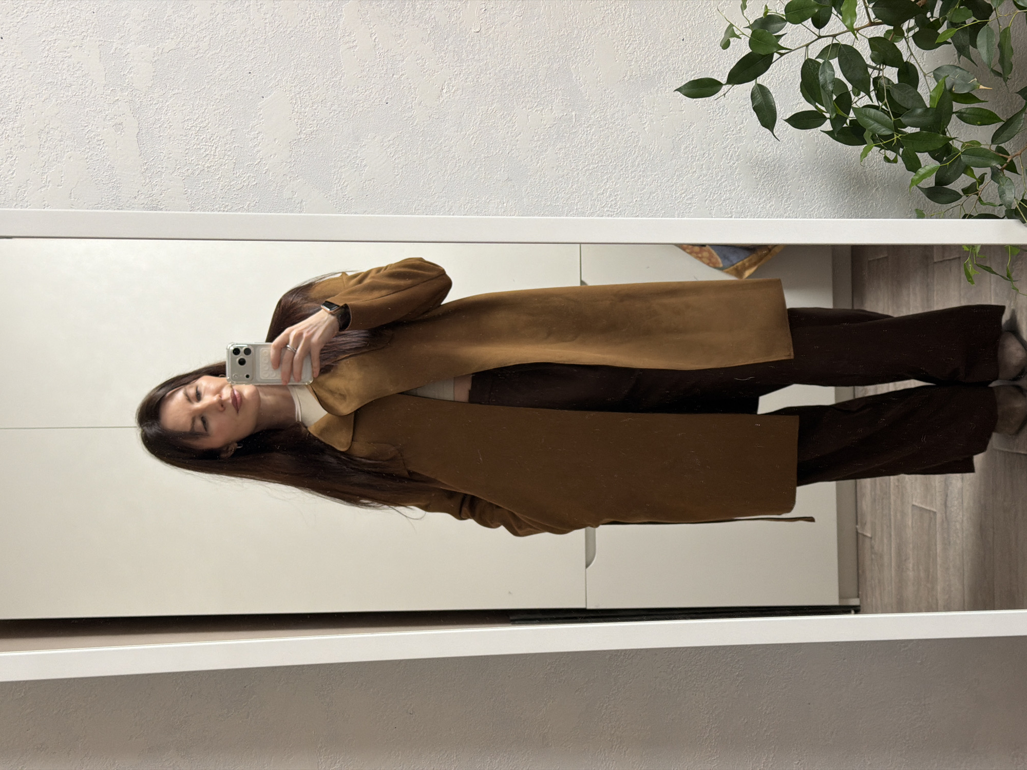 A woman with long brown hair is taking a selfie in a mirror. She is wearing a brown coat and brown pants. The reflection shows a white wall with a plant in the upper right corner and a white cabinet in the background. The floor is wood. There is no visible evidence to confirm the location as Perm, Russia.