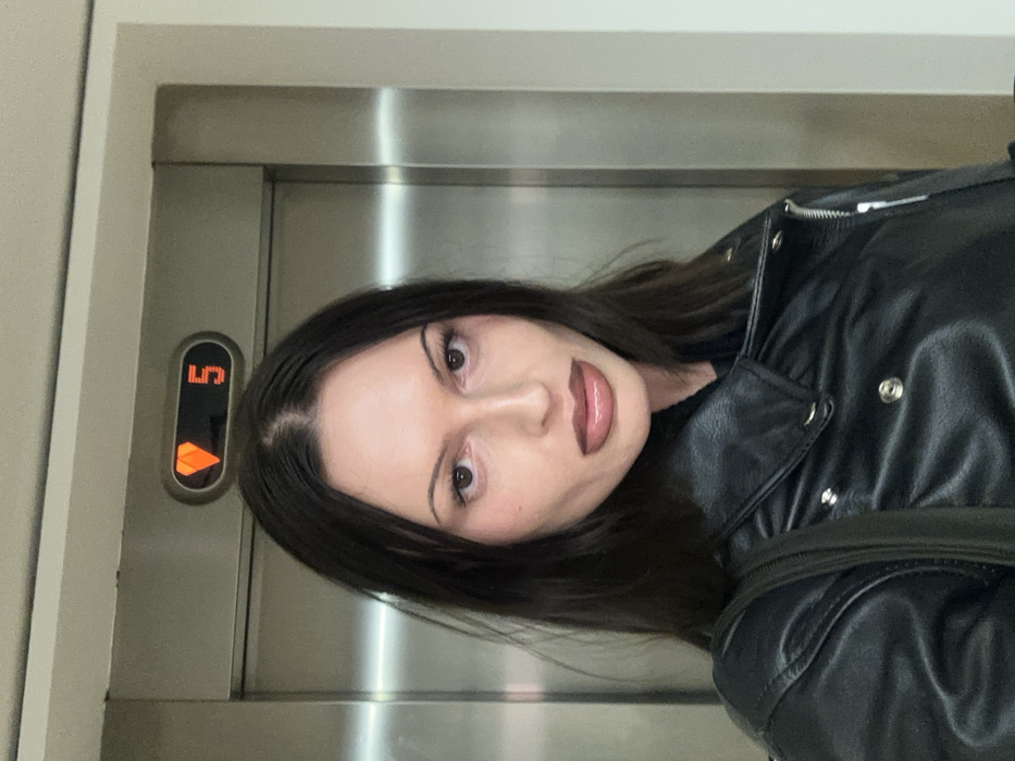 A woman in a black leather jacket stands in an elevator, facing the camera. The elevator panel shows the number five with an illuminated downward arrow. She has dark hair, light skin, and glossy lips. Her gaze is direct, and she appears to be taking a selfie in the mirrored elevator. The scene is brightly lit with the metallic interior of the elevator as a backdrop.