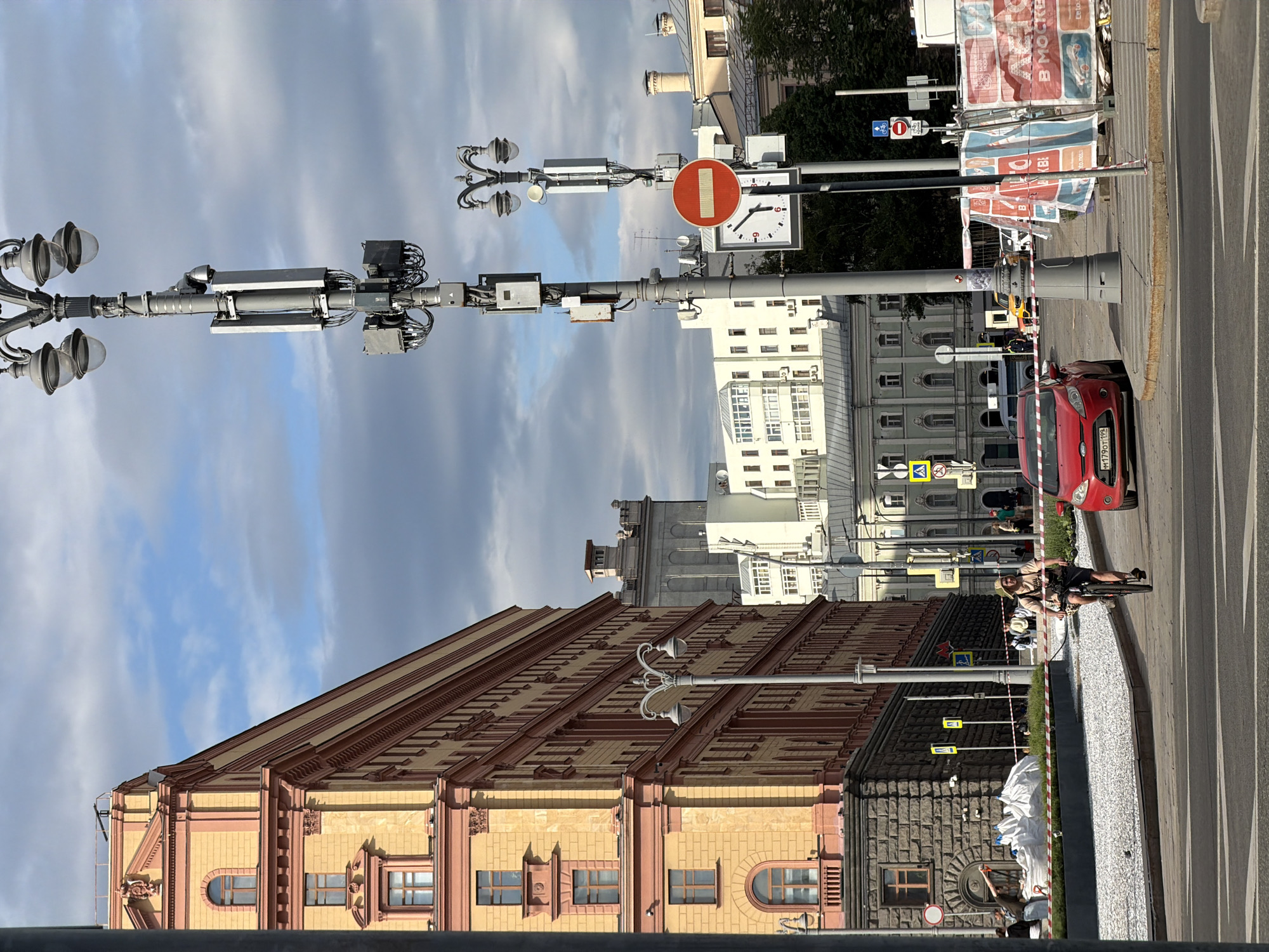 This image shows a street in Moscow, Russia. A person rides a bicycle in the street. A red car is parked on the side of the road. Buildings are visible, including one with an elaborate roof and another with a tower. A street light with numerous lights and electronic equipment is present. A "Do Not Enter" sign and a clock are attached to a pole.