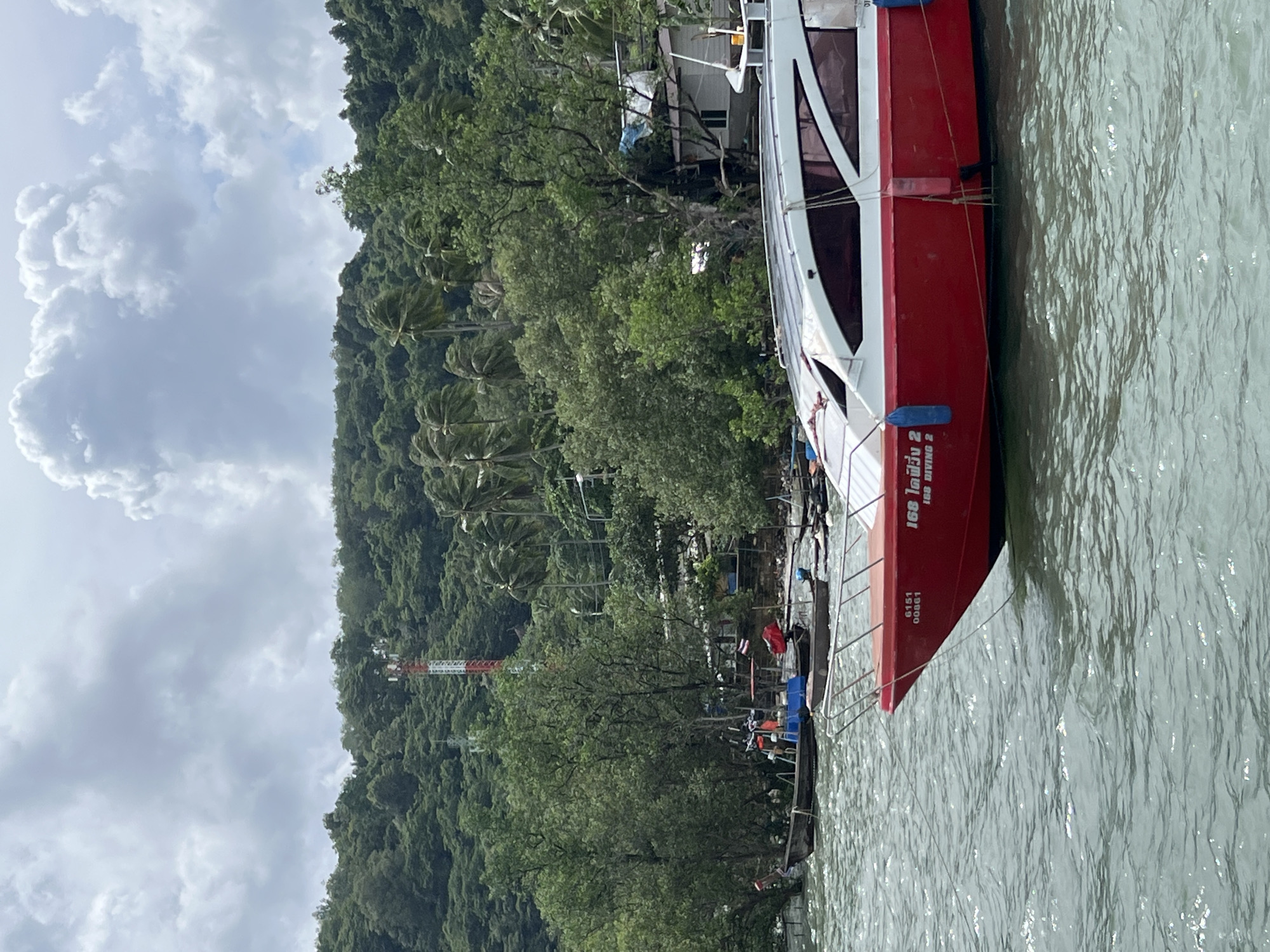 A wide shot shows a red and white speedboat docked in a body of water in Pa Khlok, Thailand. The boat has the text "6151 00861" and "ICE DIVING 2" printed on its side. The background features a dense, green, forested hillside with scattered palm trees, several buildings, and a red and white radio tower. The sky is overcast with white, puffy clouds. Several other smaller boats are visible in the distance, as well as a few people on a dock. The water is choppy and gray-green.