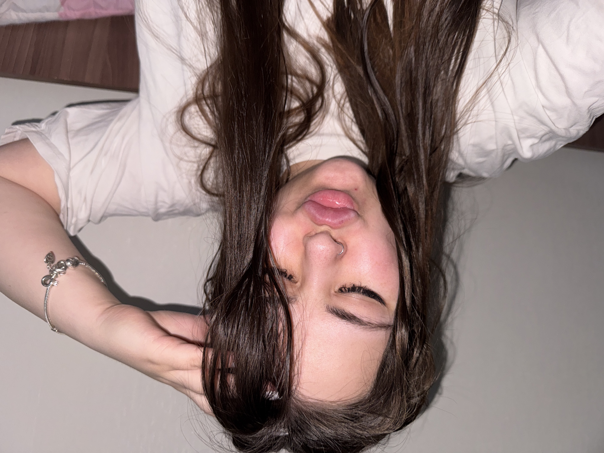 A young person with long, dark brown hair is captured in a close-up, top-down perspective, lying on their back. Their head is positioned towards the bottom of the frame, with their body extending upwards. They are wearing a light-colored (likely white or off-white) short-sleeved t-shirt.
Their eyes are closed or mostly closed, and their mouth is slightly open with their tongue playfully protruding. A silver hoop piercing is visible in their left nostril. A silver charm bracelet is worn on their left wrist, and their left hand is gently resting near their head, with fingers touching their hair.
The setting appears to be indoors, likely on a bed or a soft, light-colored surface. A dark brown wooden surface, possibly a headboard, and a glimpse of pink fabric are visible in the upper left background, suggesting a bed. The lighting is soft and artificial, indicating an indoor environment, possibly in the evening or at night. The overall mood conveys relaxation or playfulness. There is no visible text in the image.