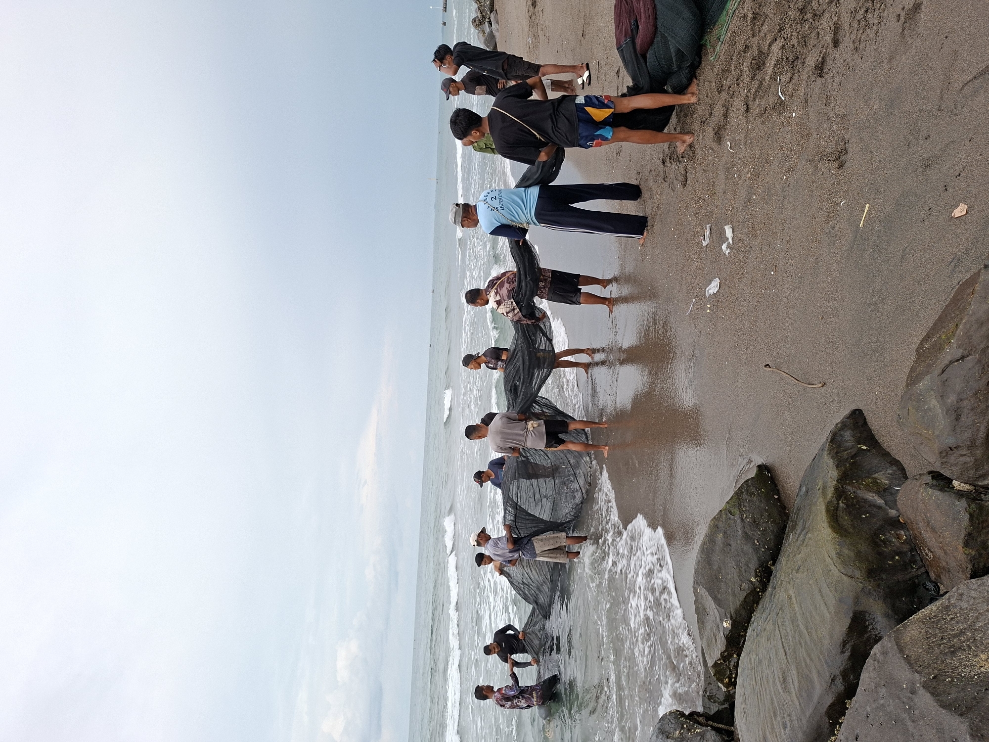 The image depicts a group of approximately ten to twelve men engaged in a communal fishing activity on a dark sandy beach in Lhokseumawe, Indonesia. The men, mostly young adults, are dressed in casual attire including t-shirts, shorts, and long pants, with several being barefoot. They are actively handling large, dark fishing nets, some standing in the shallow, breaking waves of the ocean while others are on the wet sand. The nets appear to be either being pulled ashore or prepared, indicating a traditional fishing method like seine netting.
The setting is a coastal environment with dark, coarse sand and large, dark rocks lining the right side of the beach. The ocean stretches into the distance under an overcast sky, which is light blue to grey, suggesting a cloudy day, possibly in the morning or afternoon. Small waves are breaking gently on the shore. In the far distance, faint outlines of small boats and a landmass or pier can be seen. On the sand, some small pieces of white plastic debris are visible, along with a dark, thin object resembling a rope or vine. One man in a light blue t-shirt has partially obscured text on his back, which is difficult to decipher fully but appears to include "LUHUR" and numbers. The overall scene conveys a sense of collective effort and routine work in a fishing community.