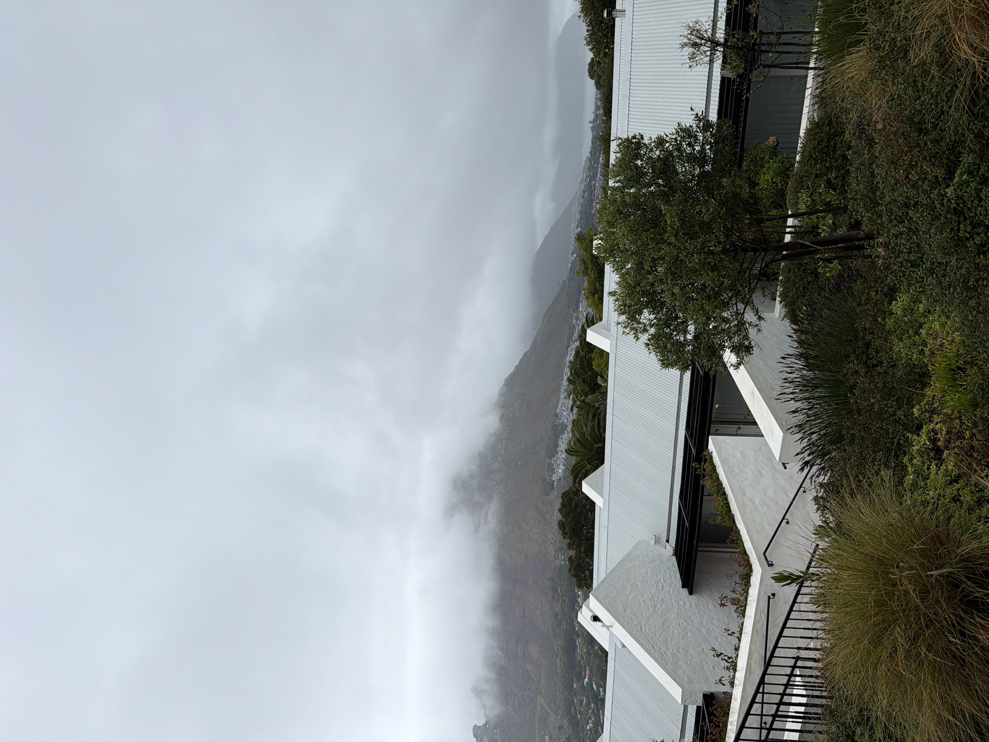 The image depicts a cloudy, overcast scene in Hout Bay, South Africa. A mountain is partially obscured by low-lying clouds. In the foreground, there are modern white buildings with corrugated metal roofs, surrounded by greenery and small trees. A black metal staircase is also visible in the foreground. The weather appears to be misty or rainy.