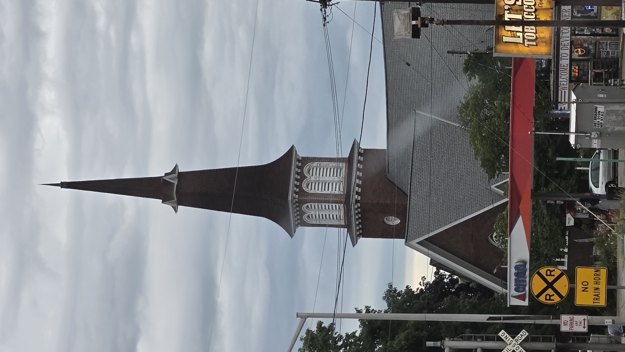 The image shows a church steeple and the roof of a church building against a cloudy sky. In the foreground, there is a Citgo gas station with a red canopy. There are also several signs visible, including one that says "NO TRAIN HORN" and another that reads "WELCOME TO DEVIL'S CORNER". A railroad crossing signal is also visible on the left side of the image. The scene appears to be in an urban or suburban setting. The location is specified as Waukesha, United States of America.