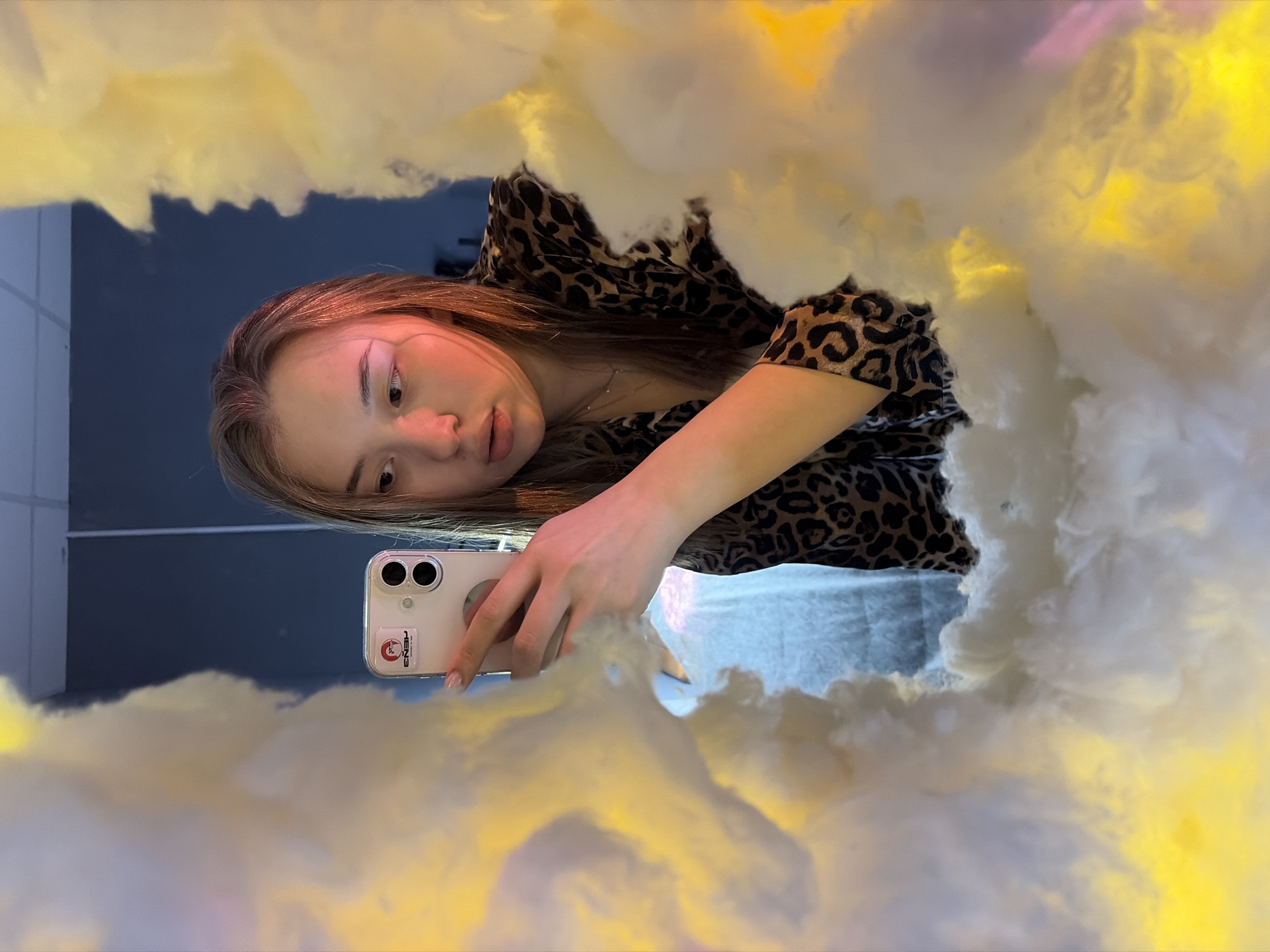 A young woman with light brown hair is taking a selfie in what appears to be a mirror or a photo booth setup. She is wearing a leopard print top. She is holding a white smartphone with two lenses and a sticker on the back. The background and foreground are filled with fluffy, cloud-like material that is lit with yellow and white light. A dark blue wall is visible in the background. Based on geotagging this was taken in the city of Chaikovsky, Russia.