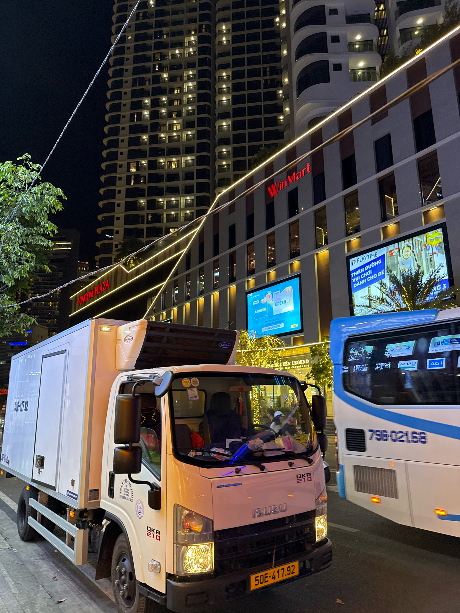 In Nha Trang, Vietnam, at night, a white box truck is parked on a city street in front of a modern building complex. The building features a large "VINCOM PLAZA" sign and a "WinMart" sign illuminated by red lettering. Several illuminated signs are also visible on the building's facade. A blue and white bus is parked next to the truck. The truck's license plate reads "50E-417.92". The bus's license plate is partially visible and reads "79B-021.6B". The street is lit by artificial lights and the illuminated building facade. Trees and foliage are visible along the street. The sky is dark, indicating nighttime.