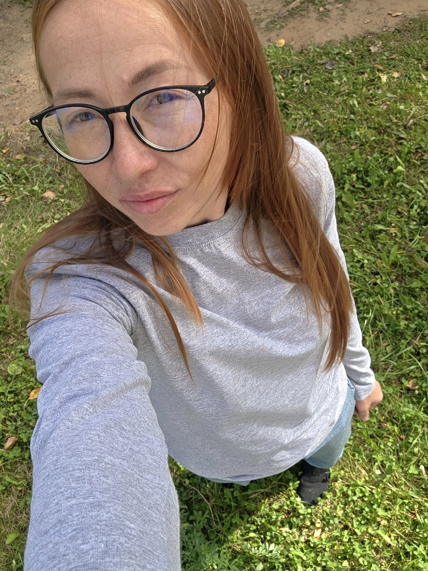 A woman with long, reddish-brown hair and freckles is taking a selfie outdoors in what appears to be a park or grassy area. She is wearing black-rimmed glasses and a light gray long-sleeved shirt, with blue jeans visible from the waist down. Her left hand is extended, holding the phone to take the picture. The background consists of green grass, some fallen leaves, and a dirt path. The lighting suggests it is daytime. The location is stated to be in the city of Chaikovsky, Russia.