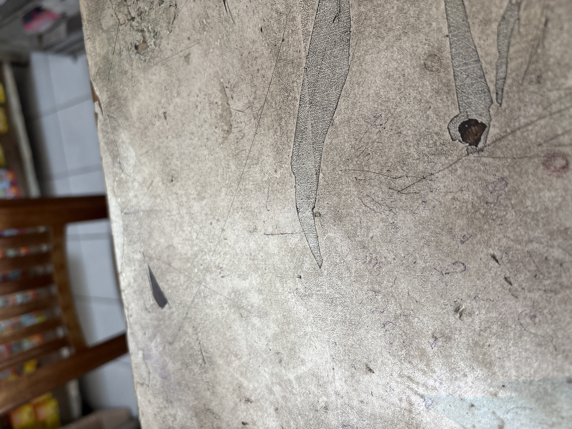 The image shows a close-up, eye-level view of a textured, weathered surface, likely a table or countertop. The surface is a light gray with visible scratches, scuffs, and some darker, irregular stains. There are also faint purple marks that appear to be scribbled or written lines. In the background, out of focus, a wooden chair with vertical slats is visible, as are shelves stocked with items, suggesting an indoor environment such as a shop or a dining area. The lighting is diffuse. There are no people or discernible actions taking place. The scene does not provide information to determine its location in Palimanan, Indonesia.