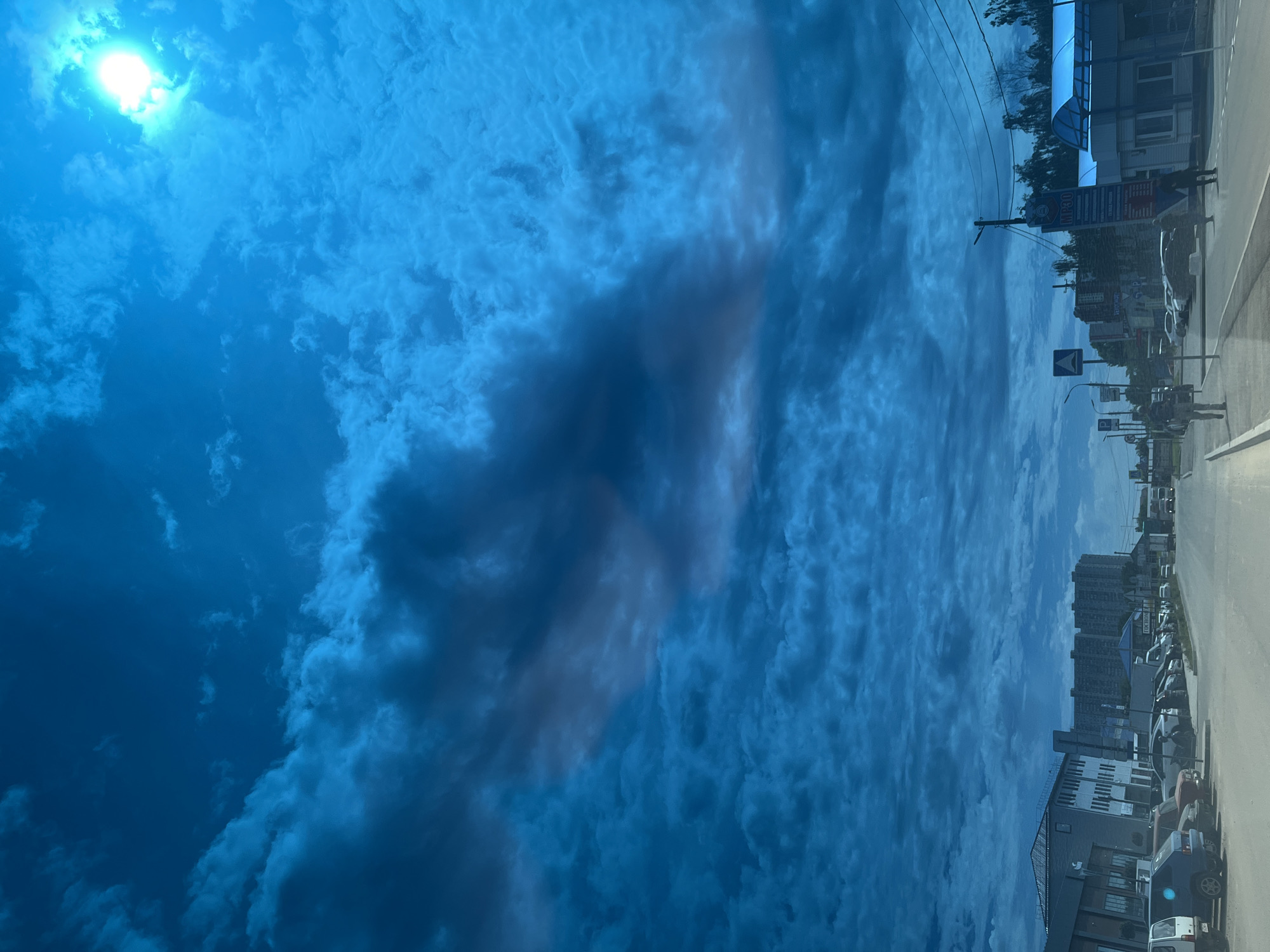 The image, taken in Krasnodar, Russia, depicts a vibrant blue sky with a street scene below, captured during daytime. The sun is visible and bright in the upper left corner, casting a strong light. The sky is filled with a mixture of bright white, fluffy clouds and larger, darker grey cloud formations, particularly prominent towards the center of the sky.
Below the sky, a road or street stretches horizontally, with buildings lining one side. These structures include a variety of commercial properties, some with awnings, and multi-story residential buildings further in the background. Several vehicles are parked along the street, and others appear to be moving.
Street infrastructure such as utility poles with overhead power lines and various road signs are visible. One notable blue sign prominently displays the text "MP-30" in white lettering. Other signs feature Cyrillic script, though the specific details are not clearly legible due to distance and resolution. A few indistinct human figures can be seen on the sidewalks, indicating general pedestrian activity. The overall image has a strong blue tint, contributing to its distinctive appearance.
