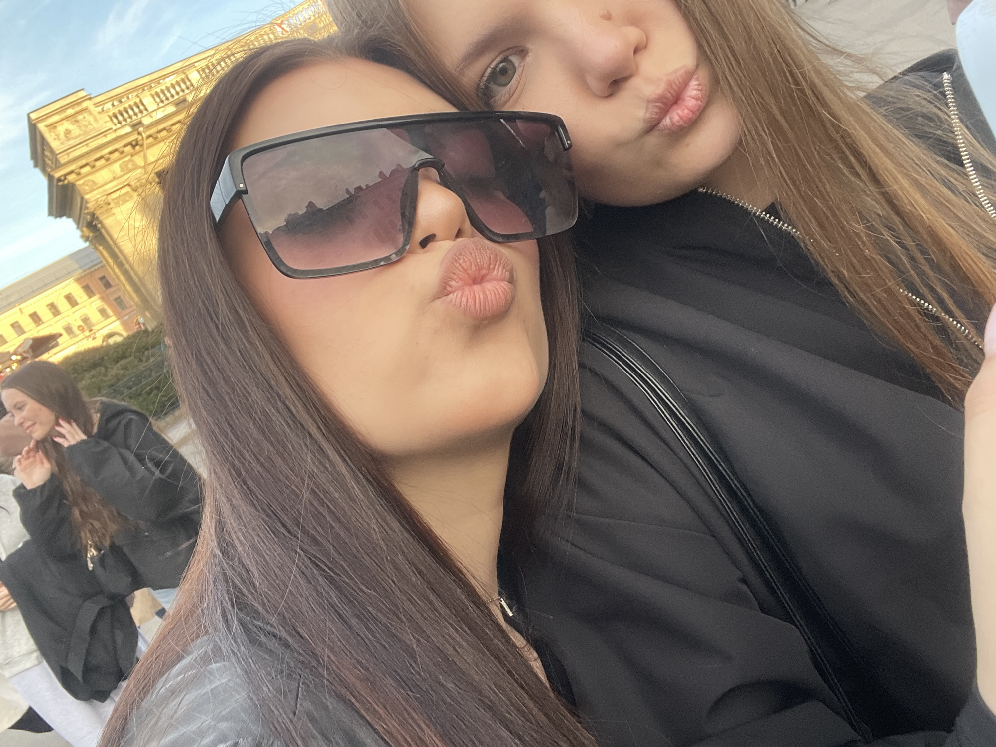This close-up outdoor selfie features two young women in Saint Petersburg, Russia, captured during daylight hours under a clear sky.
In the foreground, a woman with long, dark brown hair wears large, dark rectangular sunglasses that cover a significant portion of her face. She has her lips puckered in a "duck face" pose. A delicate necklace with a small metallic bar pendant is visible around her neck. She is wearing a dark, likely black, jacket.
Behind and slightly to the right of her, another woman is partially visible, resting her head on the first woman's shoulder. She has long, light brown hair and her left eye, with a distinctive light green iris, is clearly visible. She is also making a "duck face" with puckered lips. She wears a black jacket with a zipper detail. Their expressions and proximity suggest a friendly and playful interaction.
The background reveals a magnificent, ornate classical building with warm yellow walls and intricate architectural details, illuminated by bright sunlight. The architectural style is characteristic of Saint Petersburg. Another person, a woman with long hair wearing a dark hoodie, is partially visible in the far left background, engaged in what appears to be a separate conversation or activity. The scene evokes a casual, social atmosphere in an urban setting. No text is visible in the image.