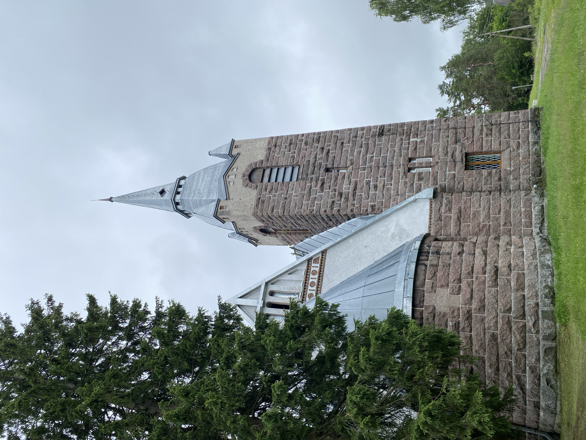 The image shows a church with a tall stone tower and a conical roof, partially obscured by a large evergreen tree on the left. The tower is constructed from rough-hewn, reddish-brown stones. It has several windows, including a tall, narrow one with horizontal slats near the top and a smaller, diamond-paned window lower down. The church has a grey, metal roof that slopes downwards. The sky is overcast with grey clouds. Green grass covers the ground in the foreground, and more trees are visible in the background. The scene appears to be in a rural or semi-rural setting.