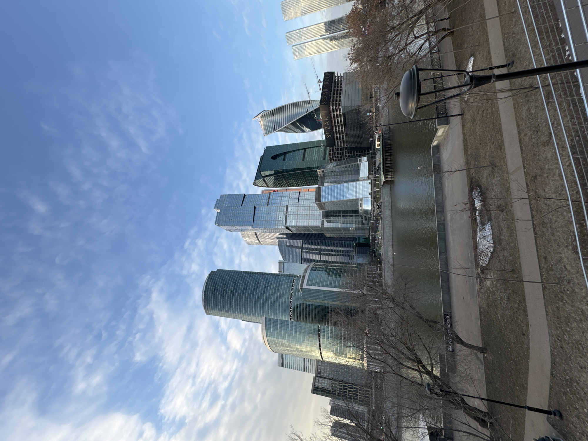 The image displays the Moscow International Business Center (Moscow City) situated along the Moskva River in Moscow, Russia. The scene is captured from an elevated perspective overlooking the river and its embankment.
The background is dominated by a cluster of modern skyscrapers of varying heights and architectural designs, primarily constructed with glass and steel, reflecting the blue sky and scattered white clouds. Notable structures include cylindrical, angular, and a distinct twisting tower. Construction cranes are visible behind some buildings, indicating ongoing development. One building under construction prominently features "SEZARGROUP" signage.
The midground features the Moskva River, with dark, calm water. A concrete embankment with a pedestrian walkway runs along the river on both sides.
The foreground consists of a park-like area along the riverbank. Bare deciduous trees with visible branches are present, suggesting a late winter or early spring season. The ground is covered with dry grass and patches of residual snow. A paved pathway curves through this area. A black, ornamental street lamp with a distinctive top design is prominently positioned on the right side, extending upwards into the frame. A metal railing, likely part of a bridge or overlook, is visible at the bottom edge. Graffiti, specifically the Cyrillic word "Кутузовский" (Kutuzovsky), is spray-painted on a concrete wall bordering the riverbank. No individuals are visible. The sky overhead is bright blue with wispy white clouds.