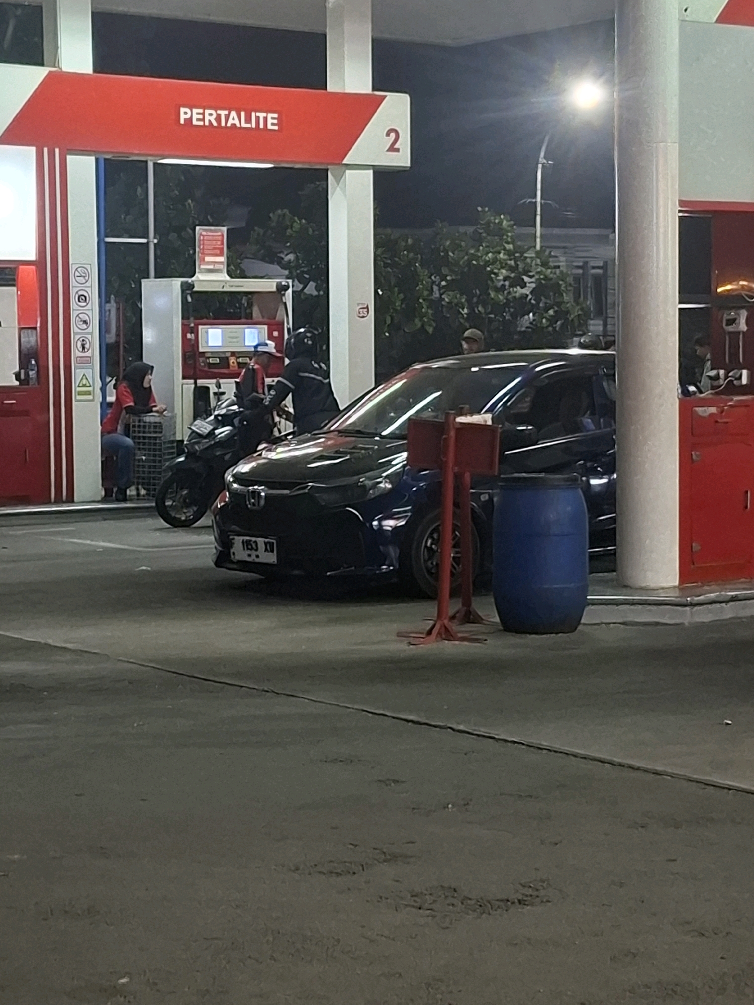 At night, at a Pertamina gas station in Karangtengah, Indonesia, a dark-colored car is parked near a gas pump. A motorcycle is also present, with a person in a helmet on it. Another person, wearing a red shirt and a headscarf, is seated on a blue plastic chair. Red gas pumps with digital displays are visible. A blue plastic barrel is positioned to the right of the car, near a red metal stand. The ground is asphalt. The scene is illuminated by artificial lighting, including a street lamp.