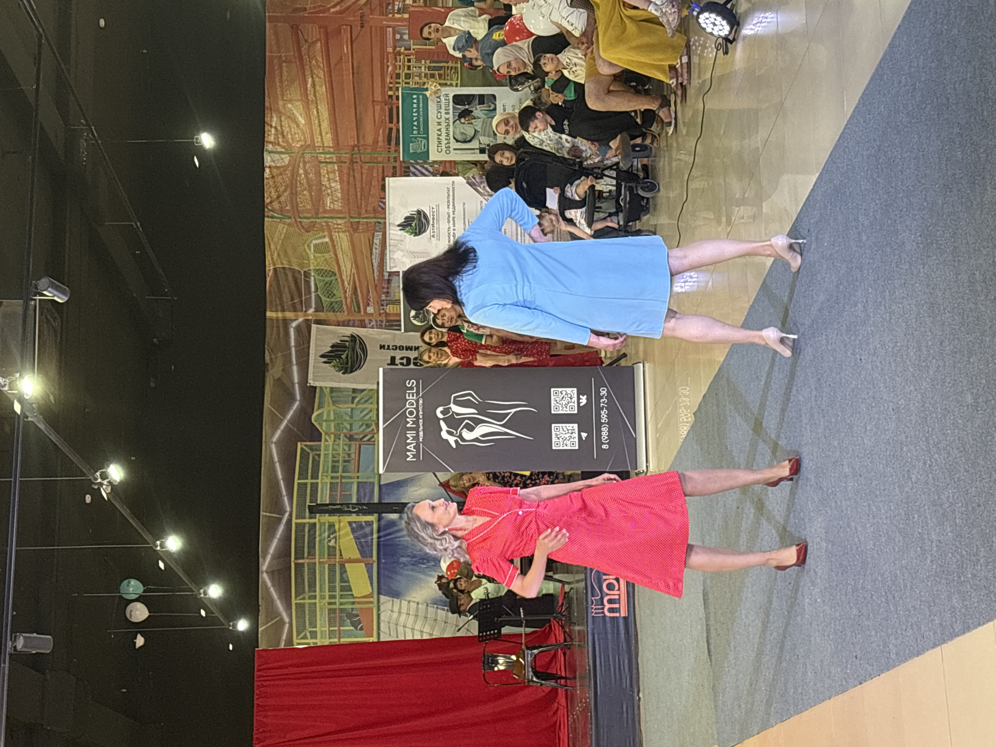 The image depicts an indoor event, possibly a fashion show or presentation, taking place at the "Mami Models" agency in Tri Potoka, Russia. Two models are central to the scene, positioned on a gray carpeted runway. The model on the left wears a bright red polka-dot dress with white trim and red high heels, while the model on the right is dressed in a light blue dress with beige high heels. Both models are facing away from the camera, looking towards an audience seated on the right side of the frame. The audience comprises men, women, and children, some of whom are watching the models. Behind the runway, a red curtain is visible on the left, and various banners and signage for "Mami Models" and other entities are displayed. The background suggests an indoor entertainment or exhibition space, with elements of playground structures visible. Lighting fixtures are suspended from the ceiling.