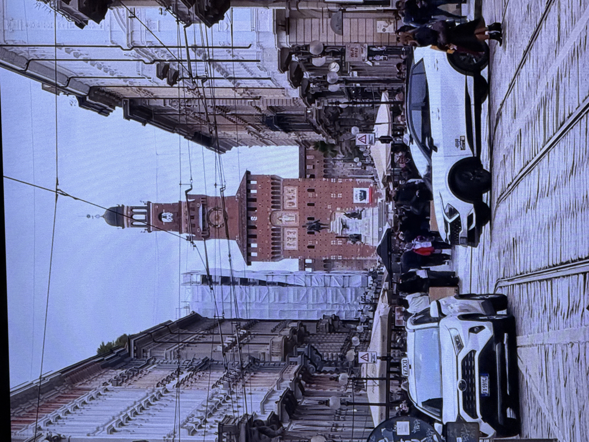 The image captures a street scene in a European city, with a prominent red brick castle or tower in the background. The street is paved with cobblestones and features tram tracks. Several buildings line the street, characterized by ornate architecture, balconies, and multiple windows. In the foreground, two white SUVs are visible, one of which is identified as a taxi with "TAXI" written on its roof. Pedestrians are scattered throughout the scene, some walking, others gathered under awnings of shops or cafes. Power lines are strung across the sky, indicating an urban environment. The overall atmosphere suggests a bustling city street on a day with overcast or diffused lighting. The image does not depict any elements suggestive of Dubai, United Arab Emirates.